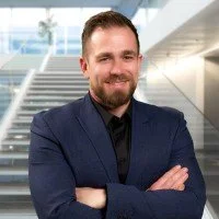 A man with a beard and short hair, dressed in a dark blue suit and black shirt, standing with arms crossed in a modern building with glass walls and stairs in the background.