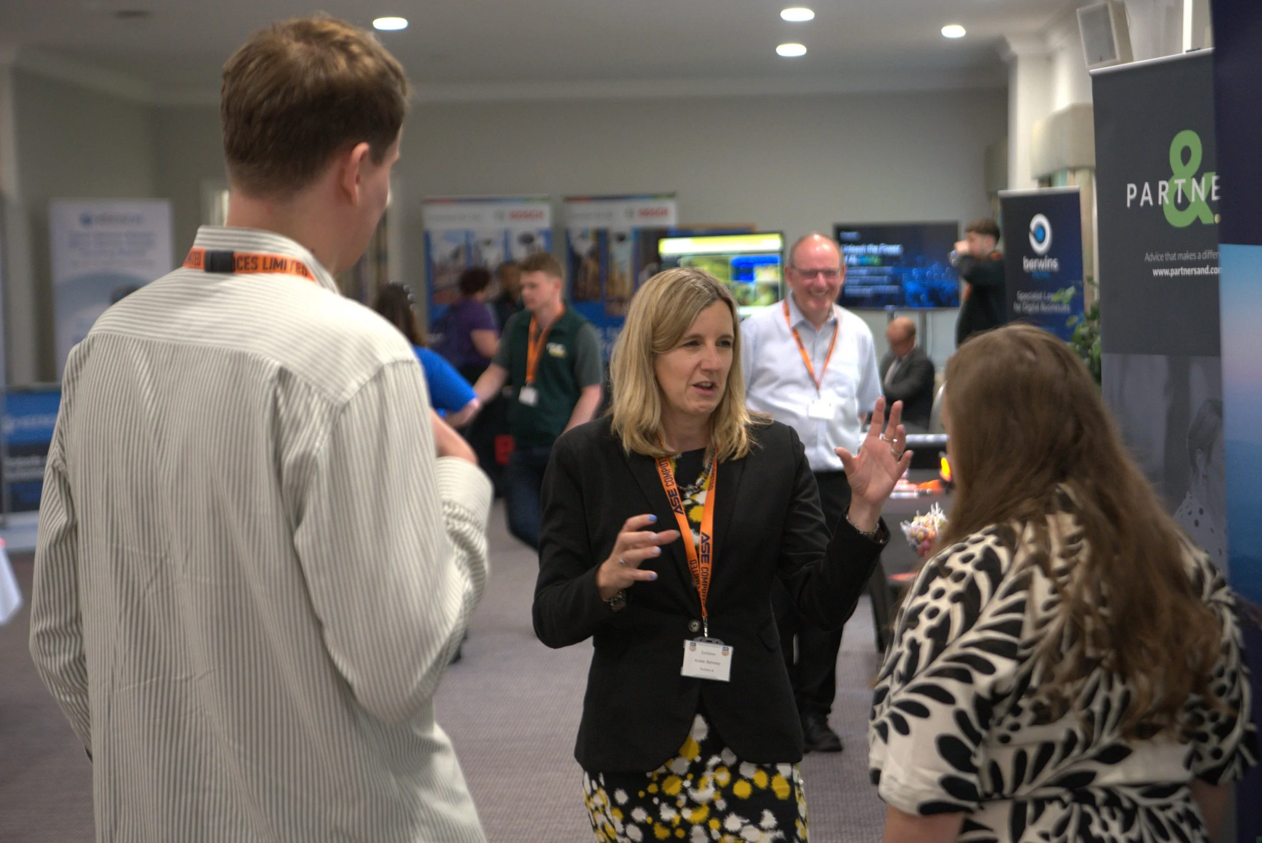 A woman in a black blazer and patterned skirt is speaking and gesturing with her hand while talking to a woman with long hair and a patterned black and white top at a conference or networking event. Several people are seen in the background, some talking and others working at booths.