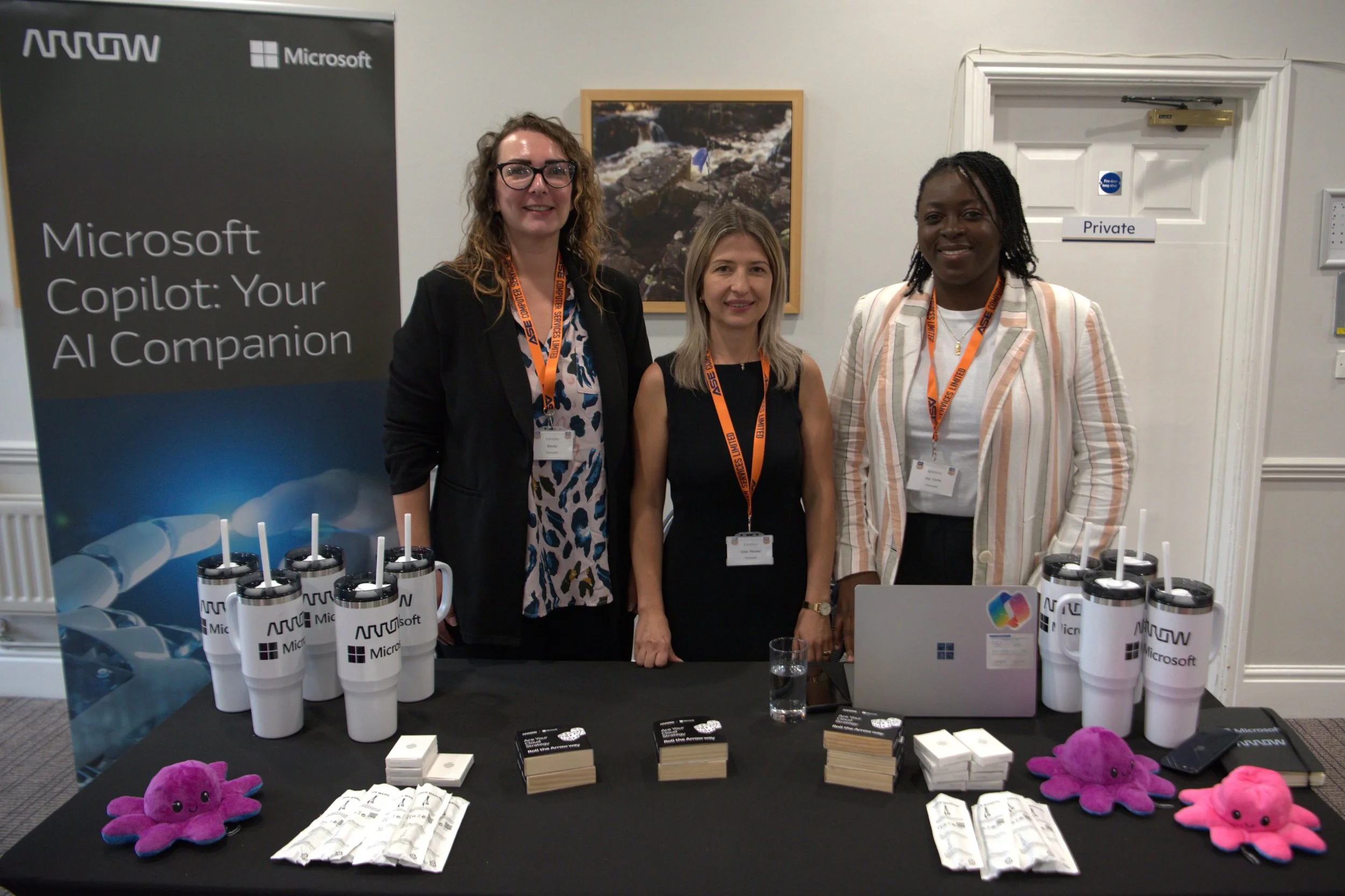 Three women standing behind a table at a Microsoft event with a large banner reading "Microsoft Copilot: Your AI Companion." The table displays white travel mugs with the Arrow logo and Microsoft branding, small plush octopus toys in pink and purple, and various pamphlets and items. The women are wearing conference badges with orange lanyards.