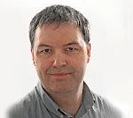 A man with short, dark hair wearing a gray shirt, smiling slightly at the camera.