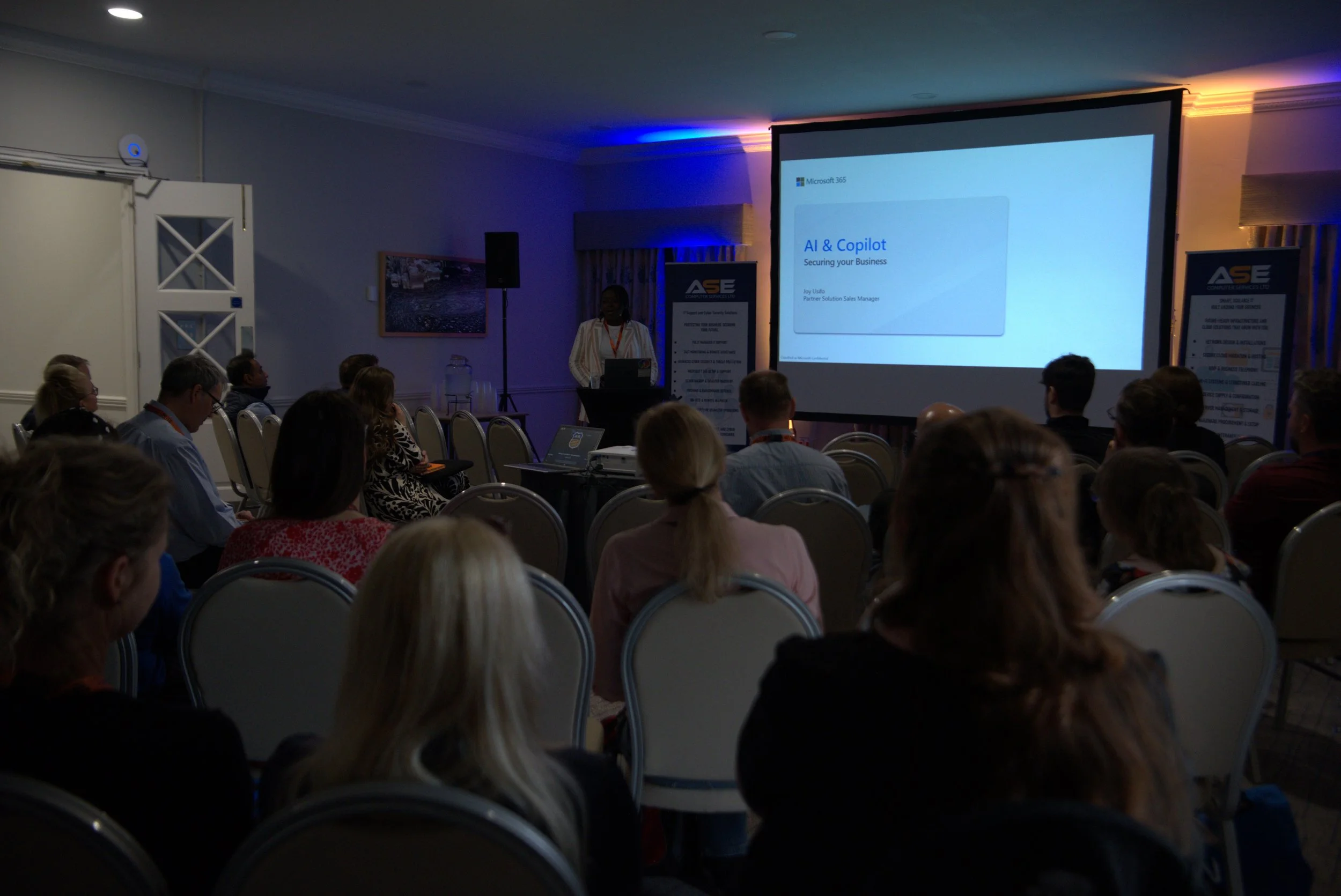 A conference room with attendees watching a presentation on AI and Copilot. The presenter stands at a podium near a large screen displaying a slide titled 'AI & Copilot'. There are banners and a projection screen in the room, with some audience members taking notes or using laptops.