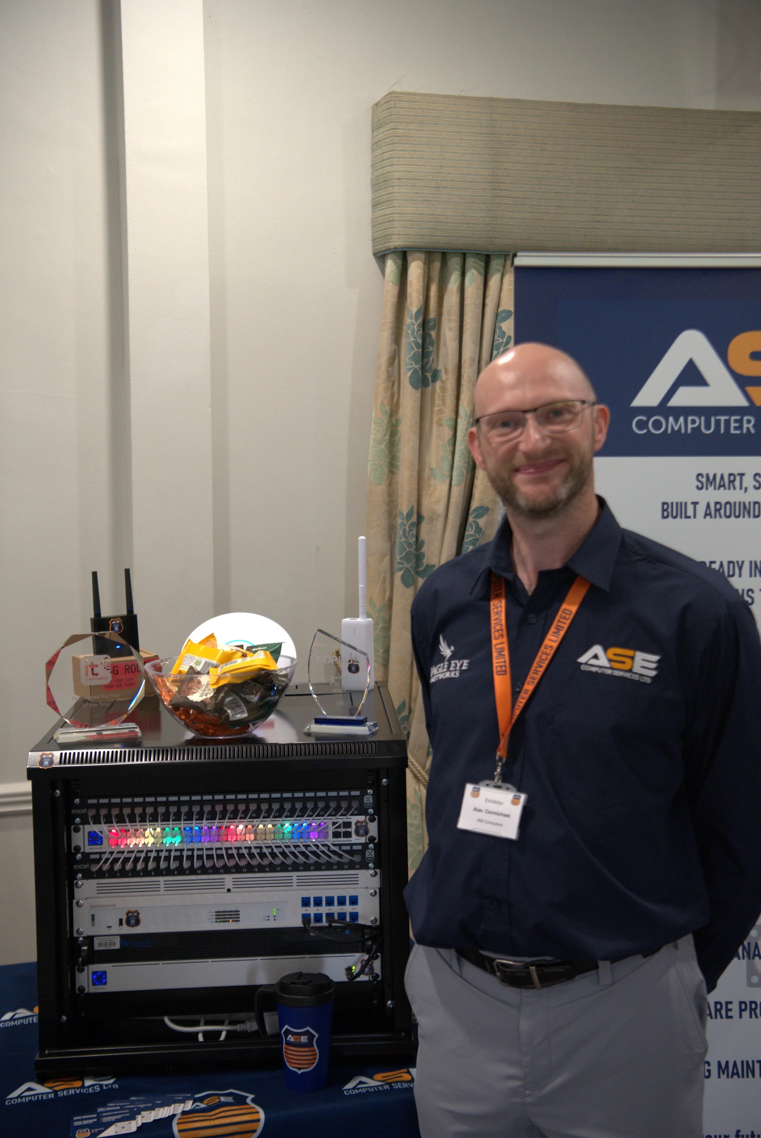 Man wearing glasses and a Navy blue shirt standing next to a table and server rack with electronic equipment, with ASE COMPUTER SERVICES banner in the background.