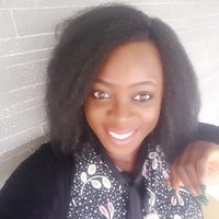 A woman with curly black hair smiling and wearing a black shirt with a white polka dot collar, standing in front of a brick wall.