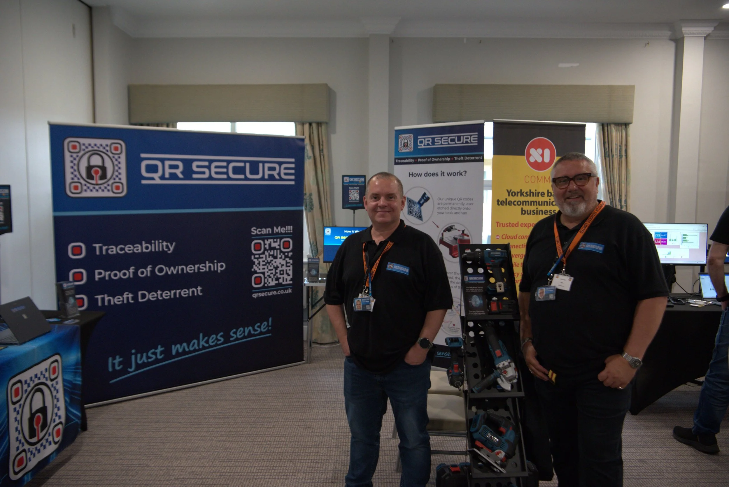 Two men standing at a booth promoting QR secure, a system for traceability, proof of ownership, and theft deterrent. The booth features banners, a display of tools, and computer monitors in the background.