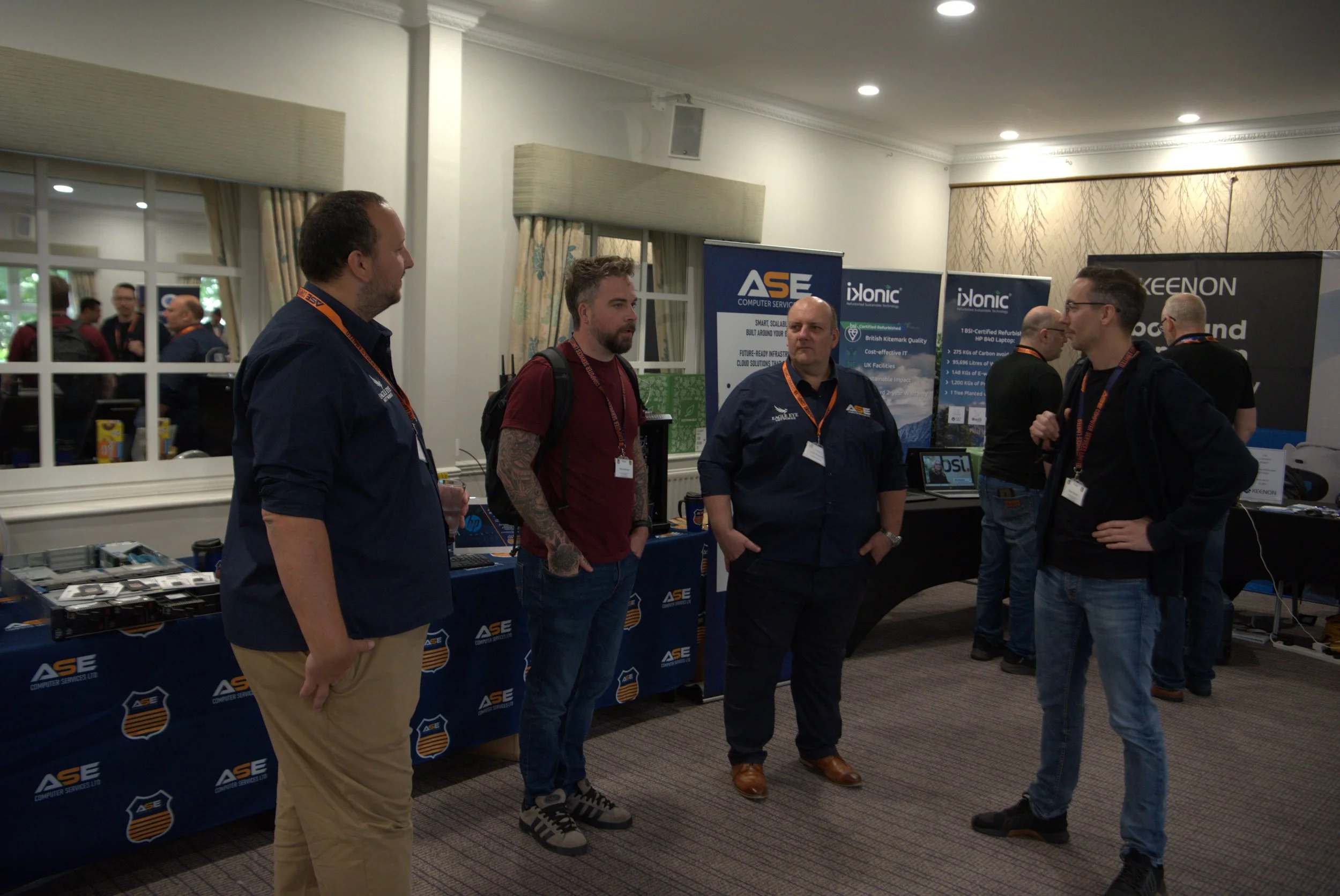 Four men are engaged in conversation at a technology exhibition. Behind them are booths with banners displaying 'ASE', 'ixonic', and 'KEENON'. The setting appears to be a conference room with a table displaying electronic equipment.