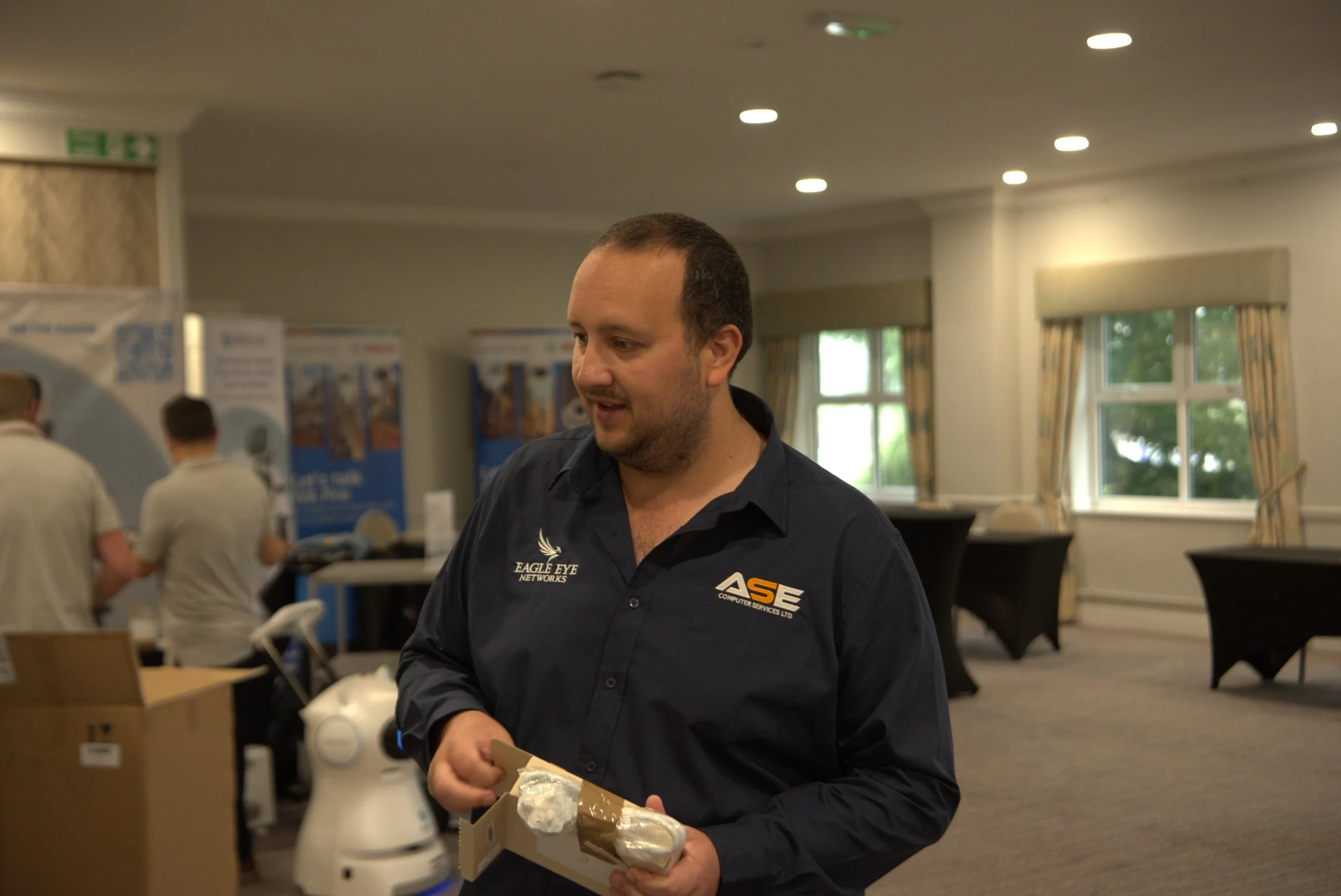 A man in a black shirt with company logos, holding a boxed item, standing in a room with other people and display banners in the background.