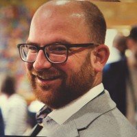 A smiling man with glasses and a beard in business attire at a social or professional event.
