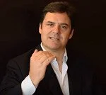 Portrait of a man in a black suit and white shirt, holding his hand near his chin and smiling.