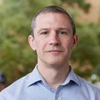 A man in a light blue button-up shirt with short brown hair, standing outdoors with green trees in the background.