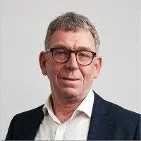 Portrait of a middle-aged man with short gray hair wearing glasses, a white shirt, and a dark blazer, standing against a plain background.