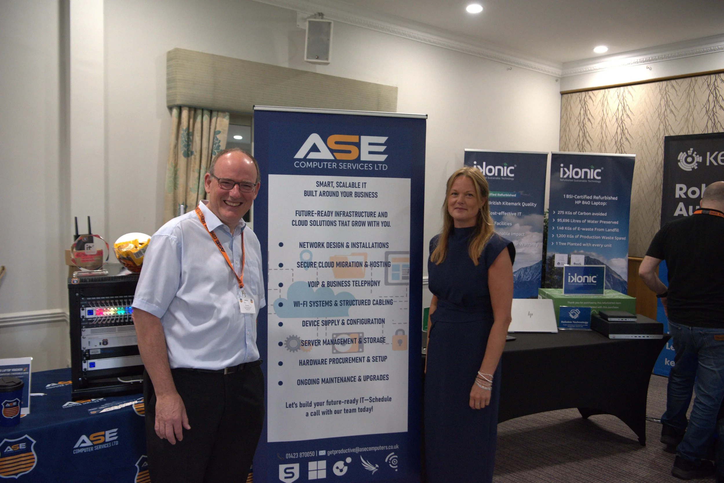 Two people standing beside a display stand at an indoor event, with banners behind them promoting IT and cloud solutions.