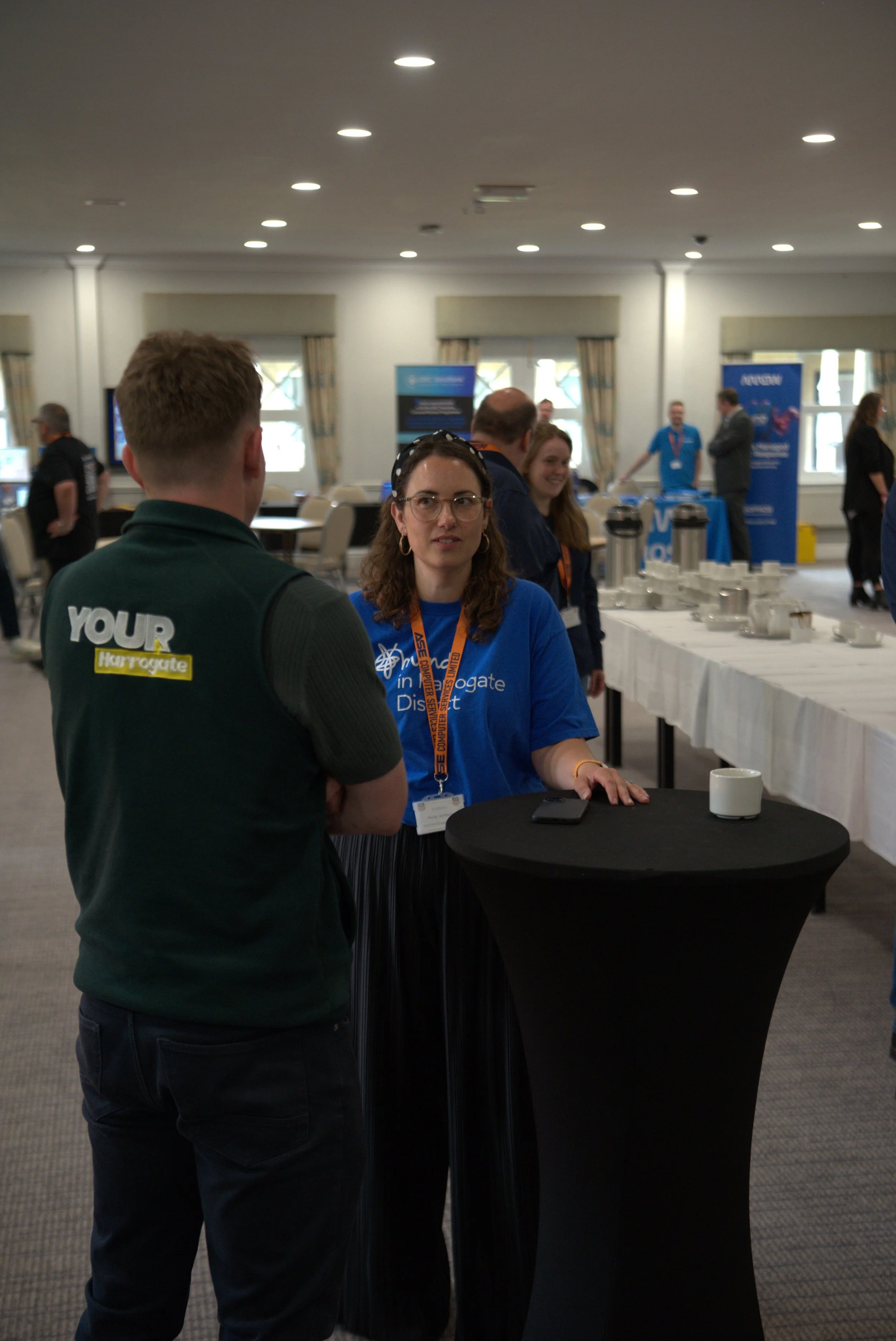 A woman with glasses and a blue shirt talking to a man with a green vest at an indoor conference or event with tables, banners, and other attendees in the background.