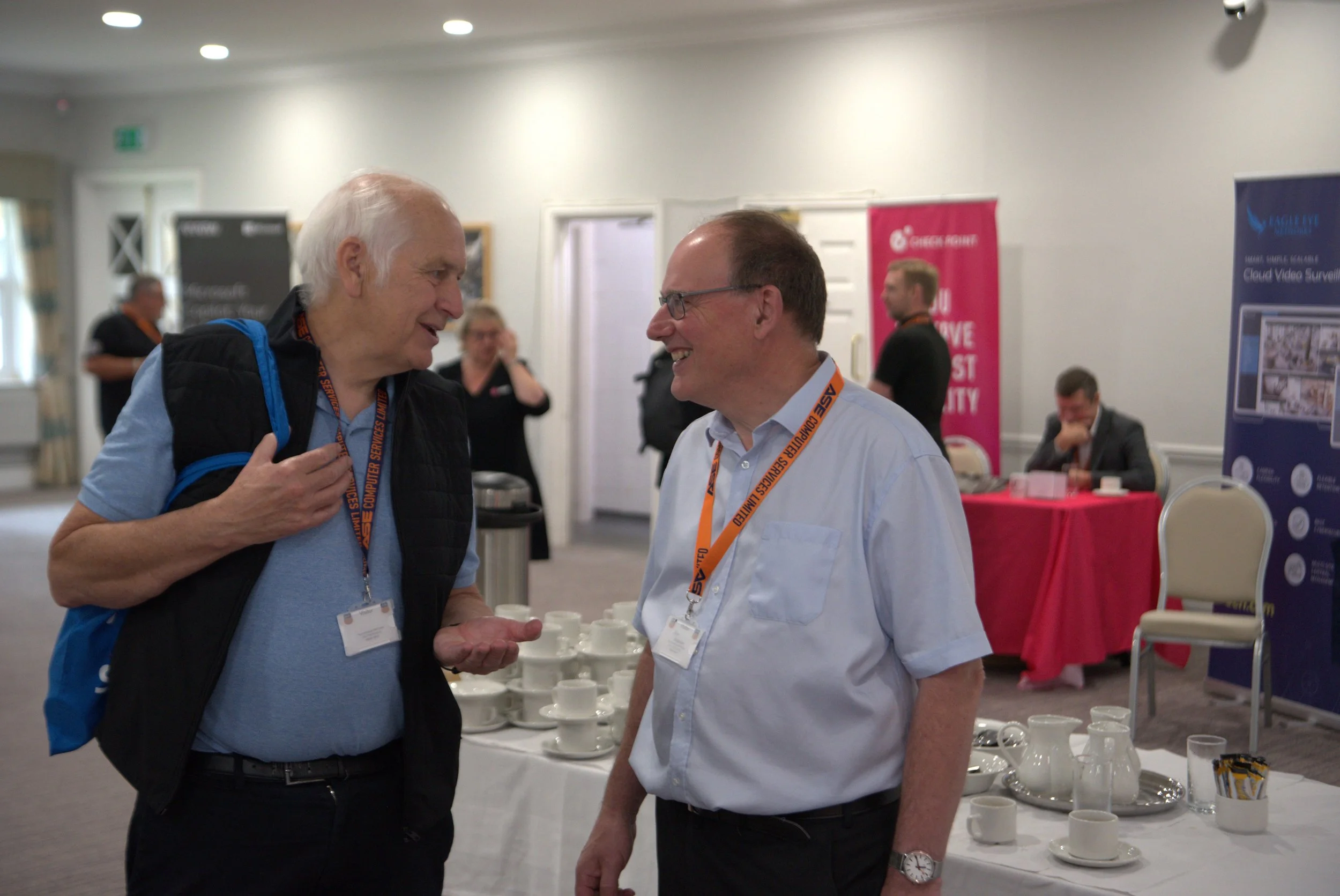 Two men are engaged in a conversation at a conference, standing in front of a table with coffee cups and tableware. There are other attendees and informational banners in the background.
