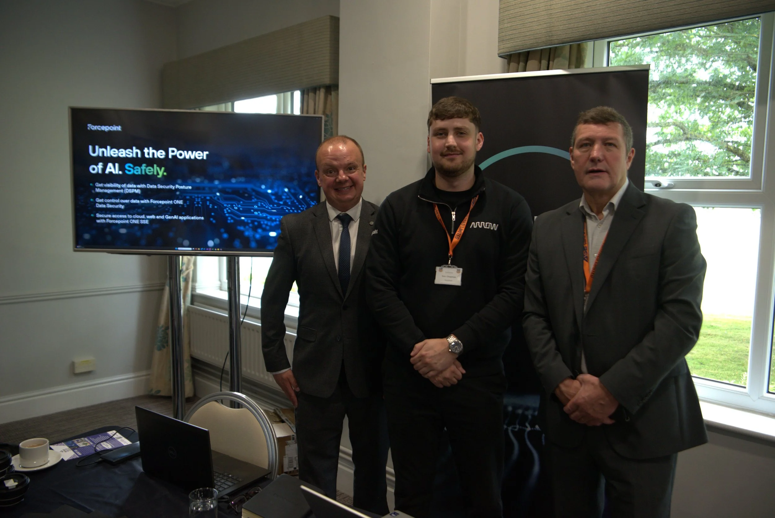 Three men standing indoors near a corporate presentation display, with a laptop and cups on a table in the foreground, and large windows showing greenery outside.