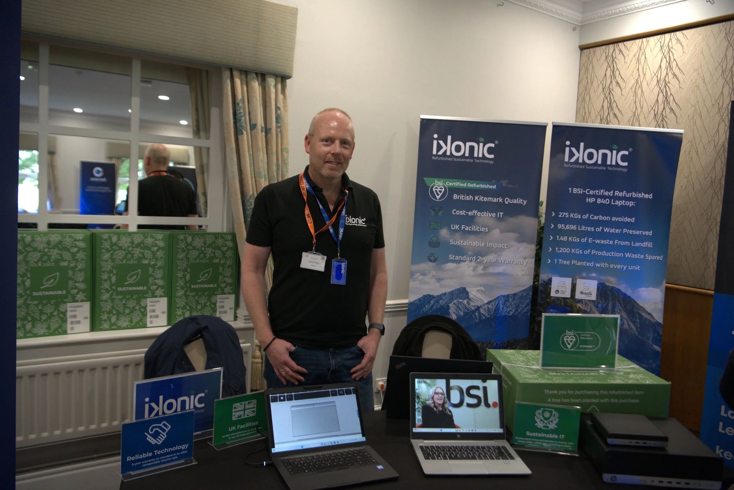 Man standing at a trade show booth for Ixonic, with banners highlighting their eco-friendly and certified refurbished technology, computers, and promotional materials on the table.