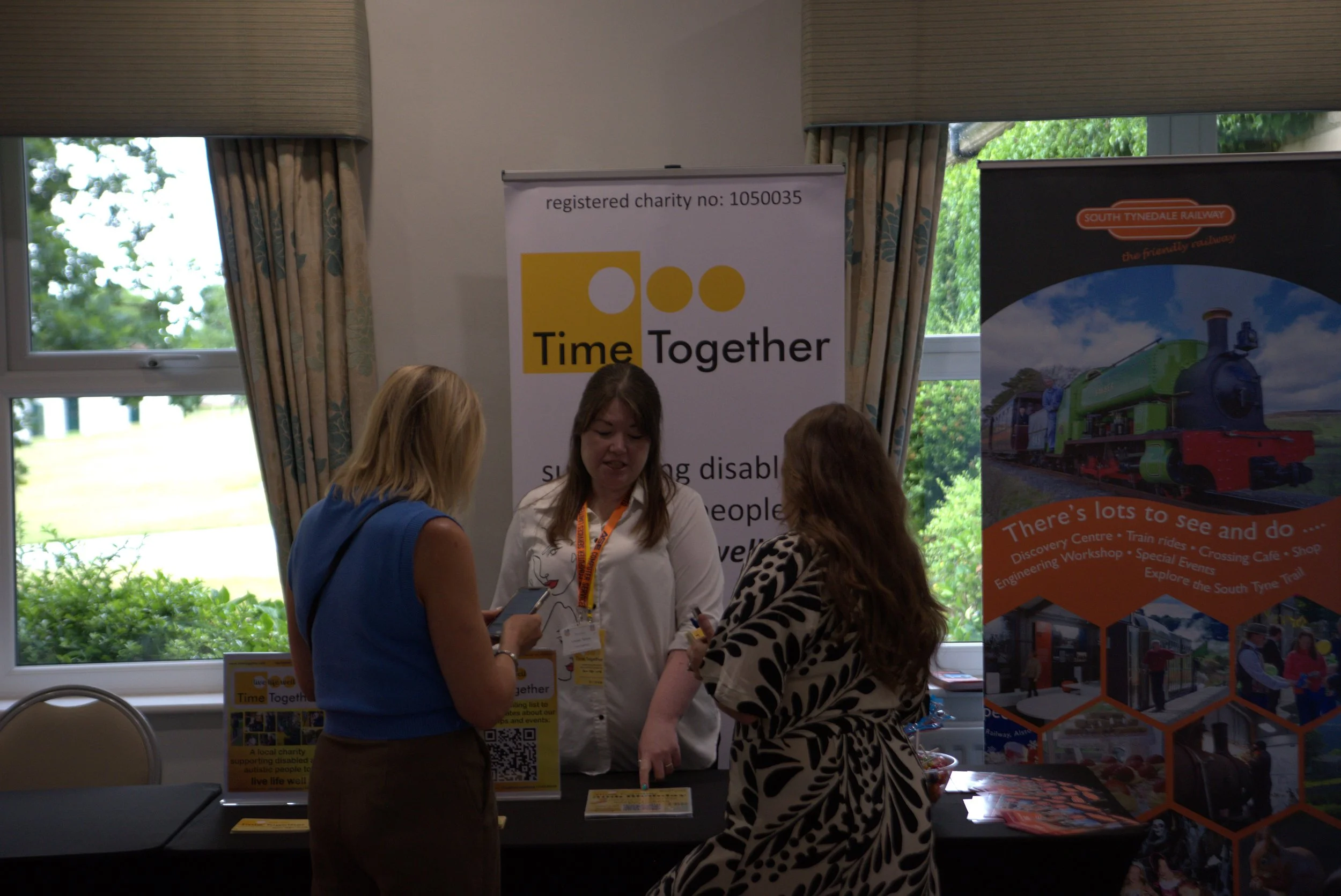 Three women at a charity event booth, with a large banner reading "Time Together" and information about supporting disabled people. The booth features promotional materials, a QR code, and a poster about South Tynedale Railway.