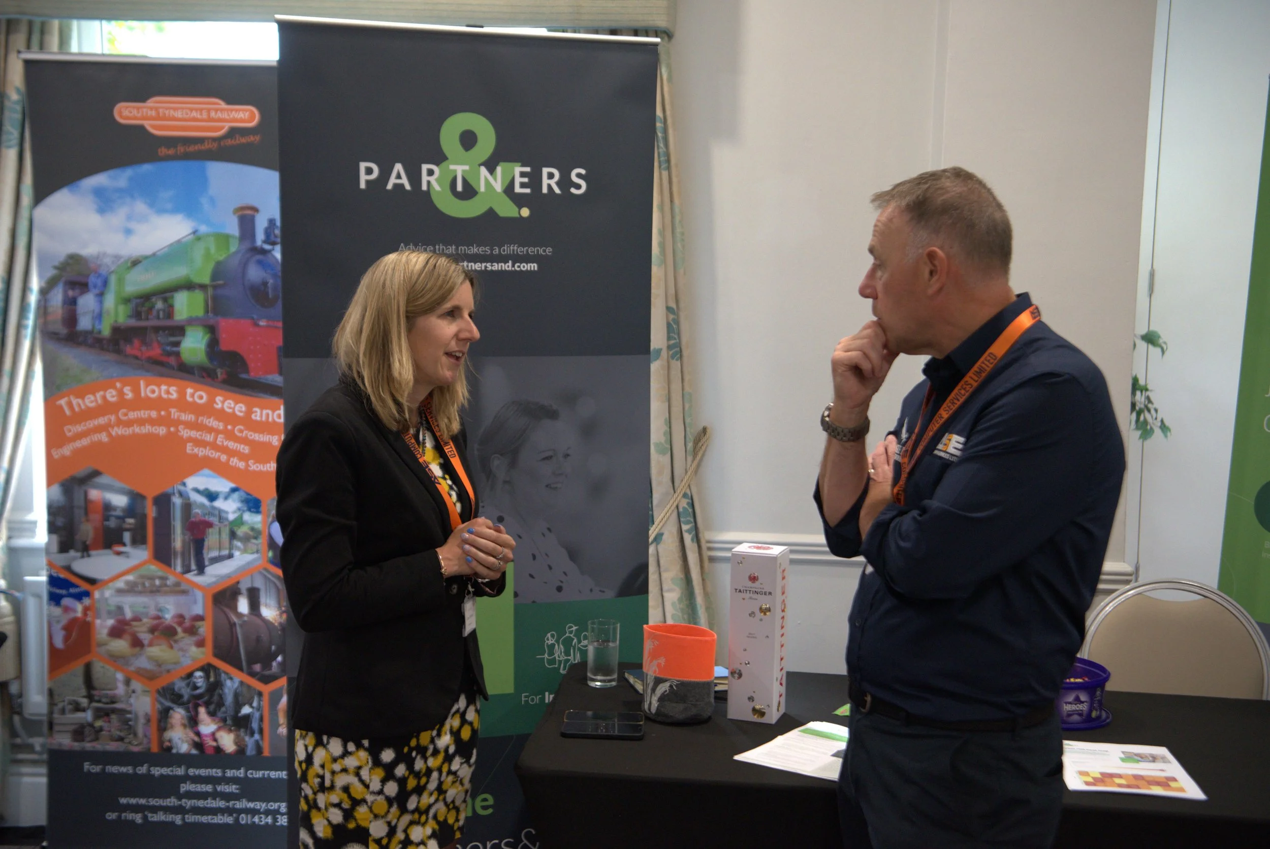 Two people in conversation at a conference booth with banners and promotional materials.