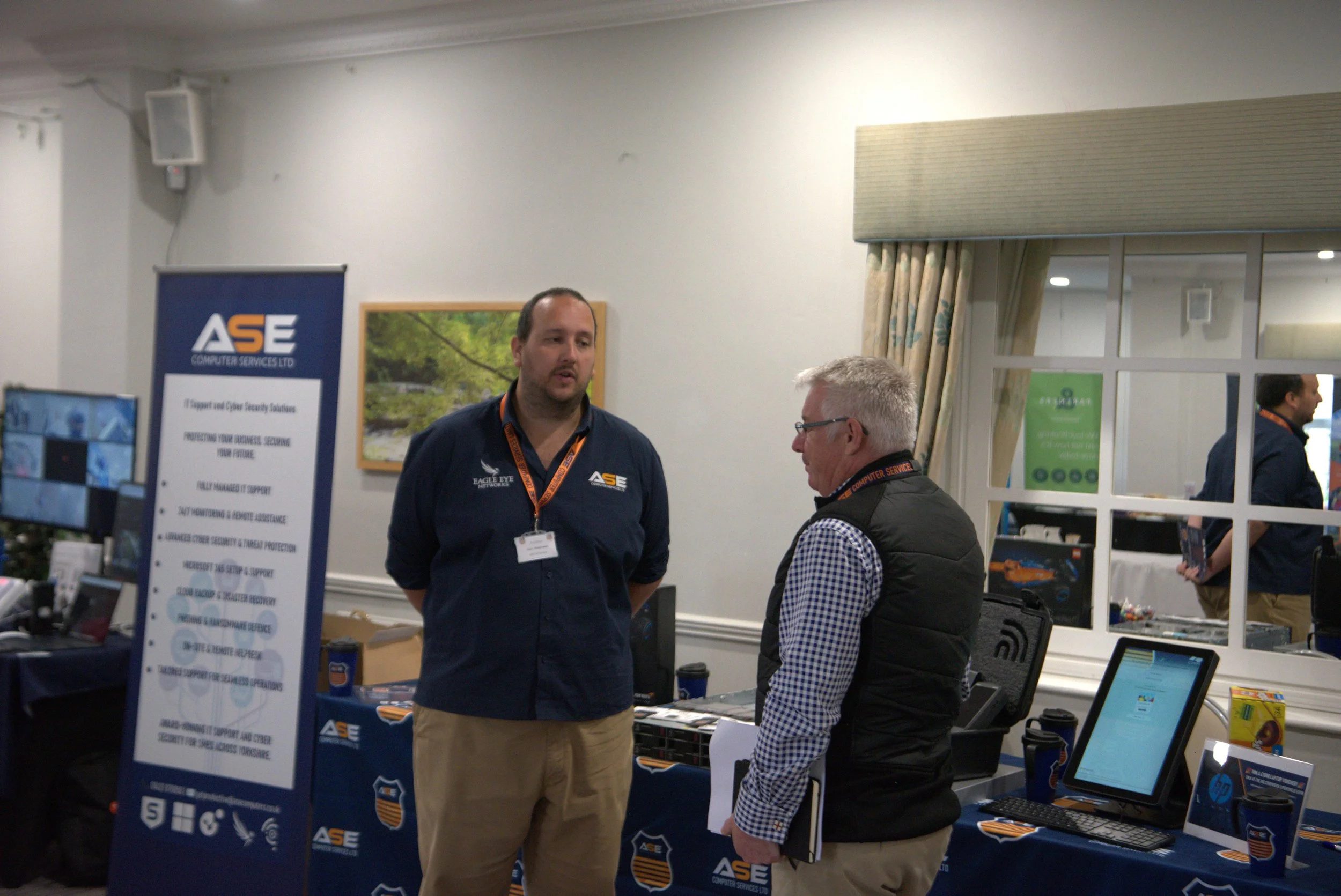Two men engaging in conversation at a technology exhibition booth for ASE Computer Services Ltd. The booth displays a banner listing services and features a computer monitor, promotional materials, and a window with curtains in the background.