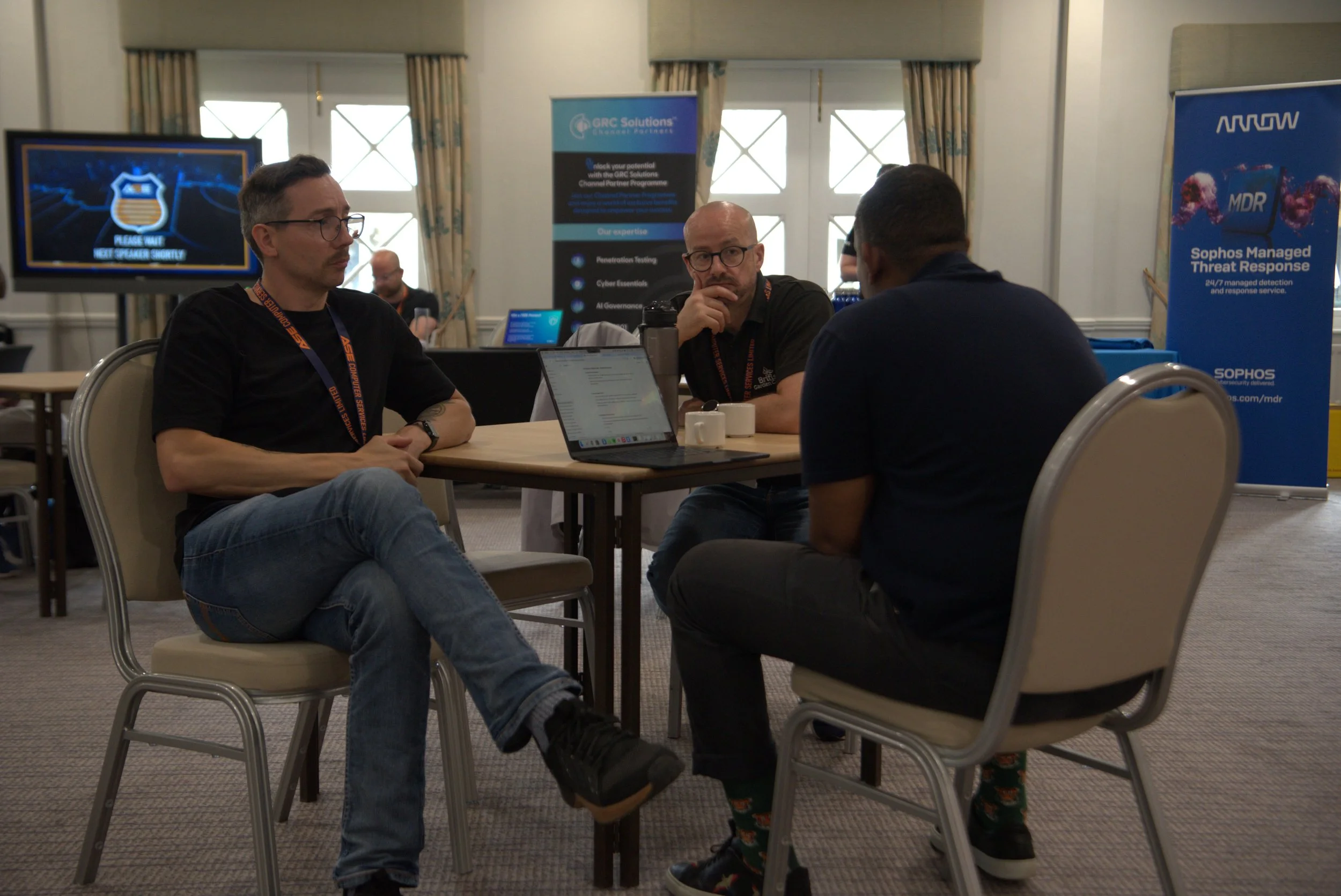 Three men sitting around a table engaged in conversation at a conference or workshop, with laptops and promotional banners in the background.