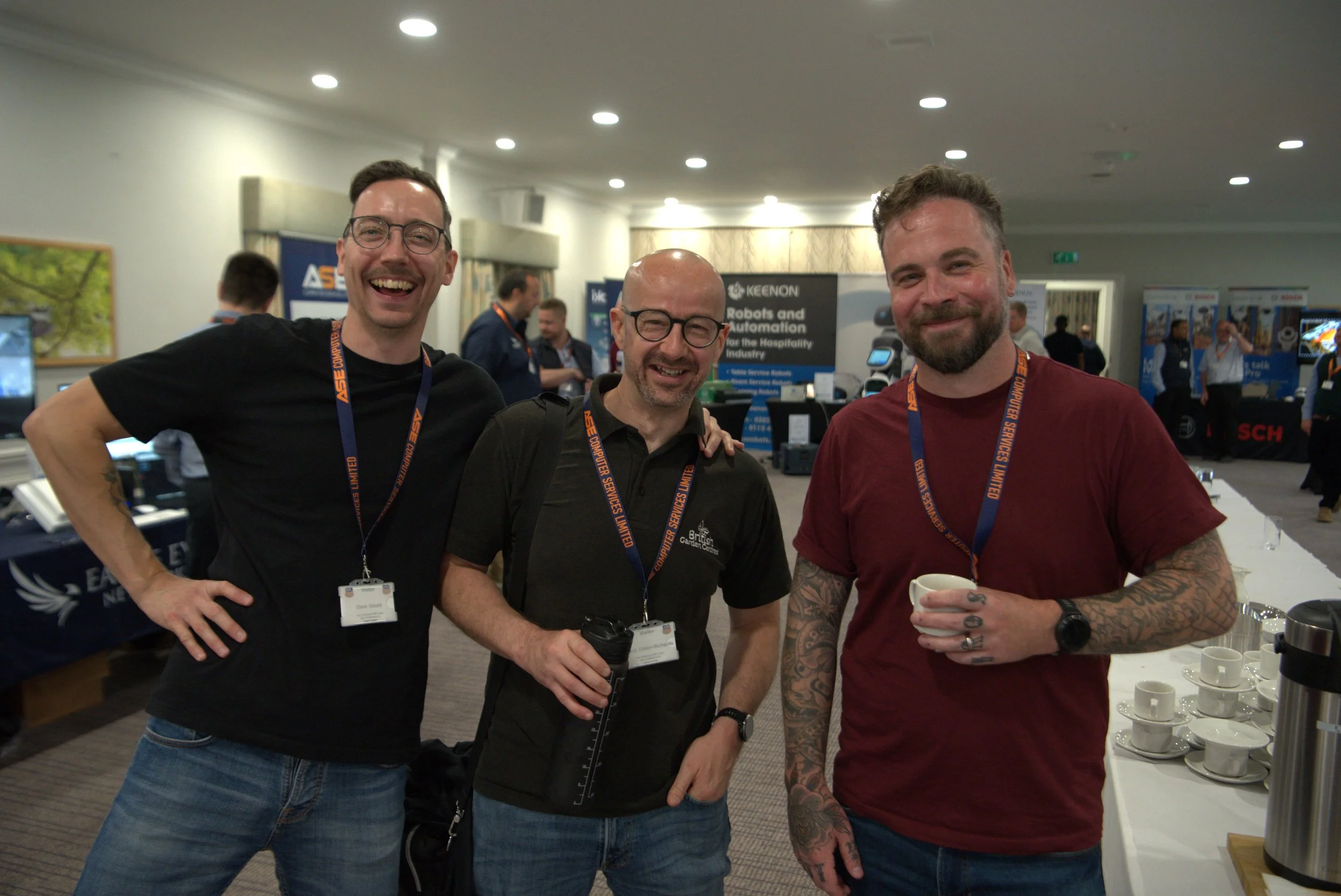 Three smiling men wearing conference badges standing together in a conference room, with booths and other attendees in the background.