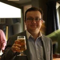 A man wearing glasses and a suit jacket holding a glass of beer in a bar or pub setting.
