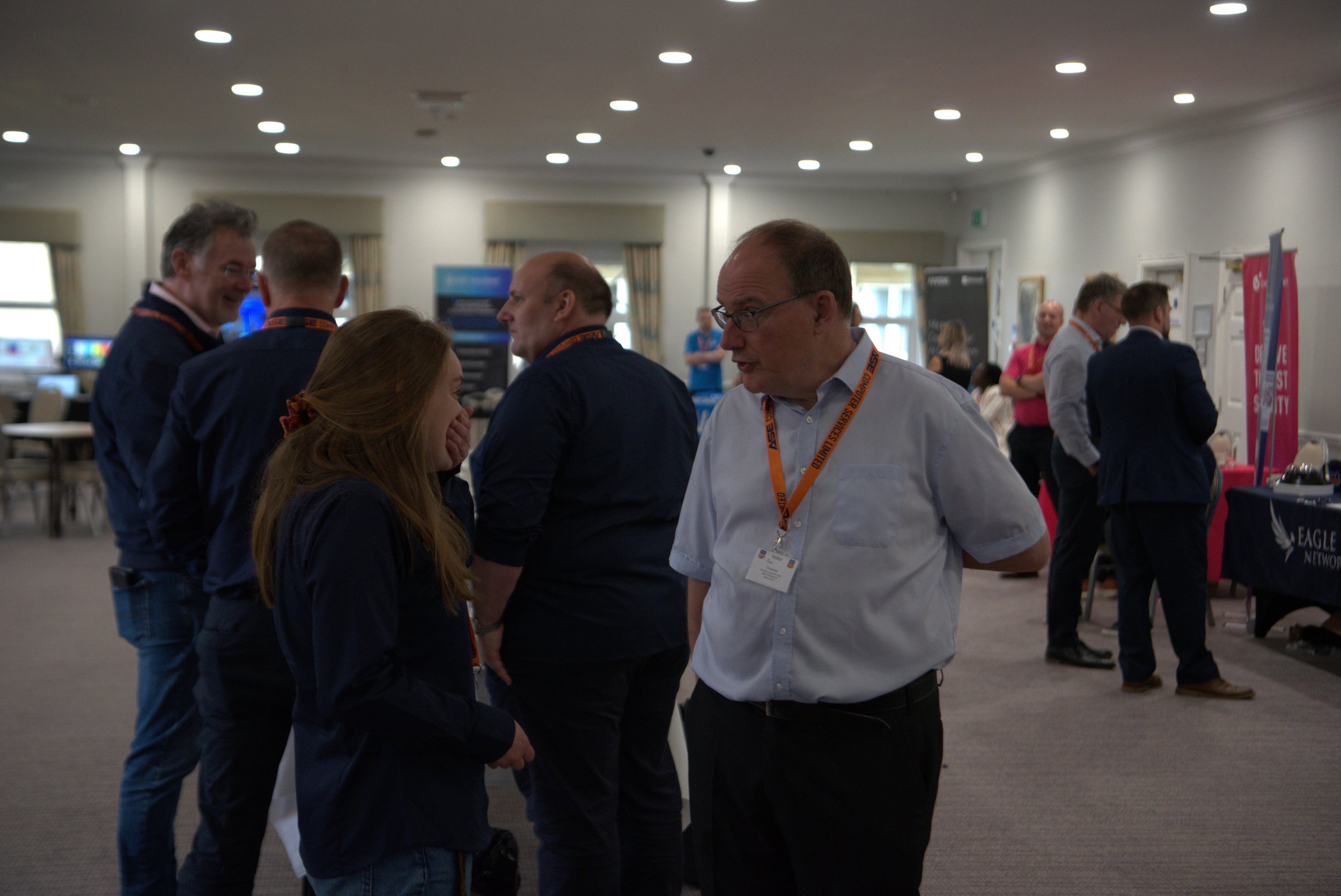 Group of people talking at a conference or exhibition in a large room with display booths and tables.