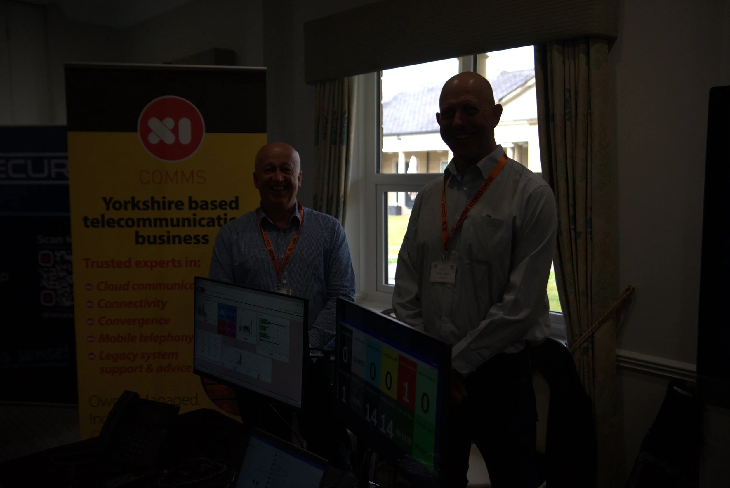 Two men in business attire standing by computer monitors during a conference or presentation, with a Yorkshire-based telecommunication business banner in the background.