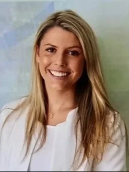 Smiling young woman with long blonde hair wearing a white top, standing against a light blue background.