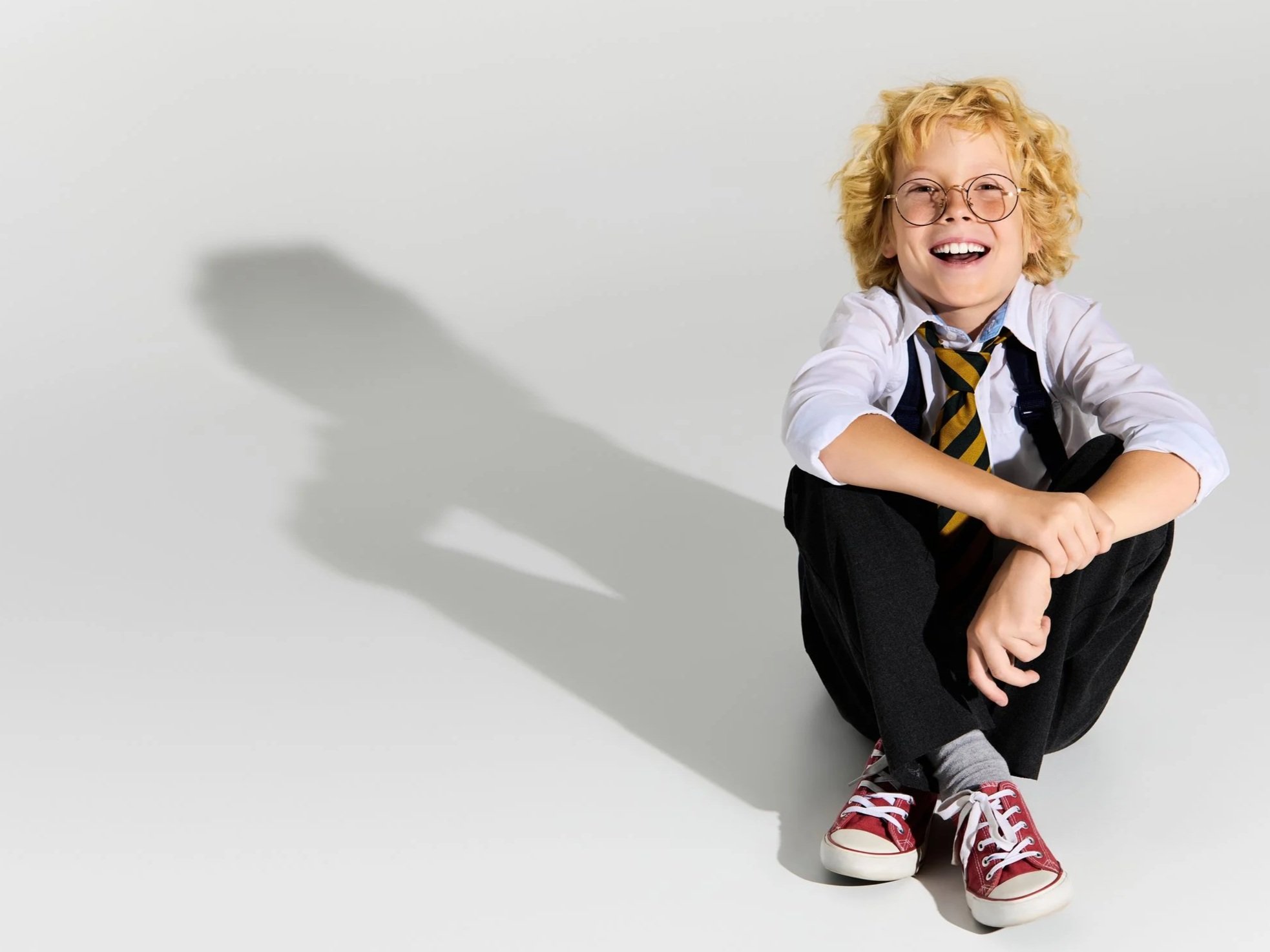 A young boy with curly blond hair wearing glasses, a white shirt, a striped tie, dark pants, gray socks, and red sneakers, sitting on the floor with a white background.