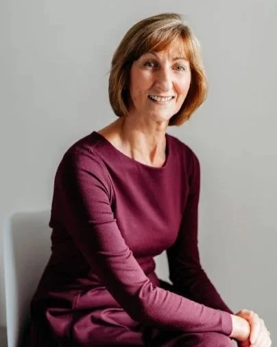 A woman with short brown hair smiling, wearing a long-sleeved maroon dress, sitting against a plain light gray background.