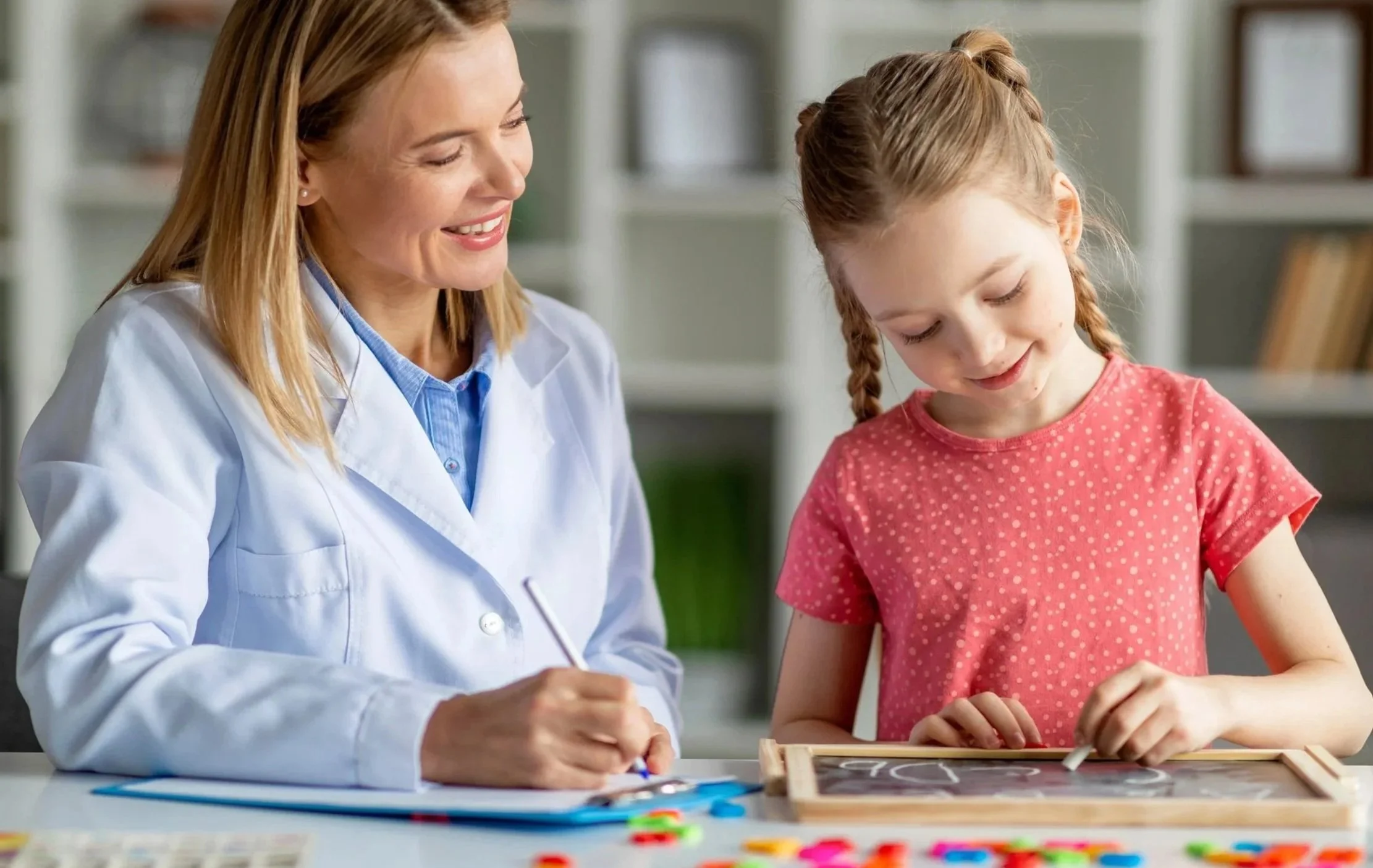 A woman in a white coat and a girl with braided hair and a pink polka-dot shirt are smiling while drawing on a chalkboard in a bright room.
