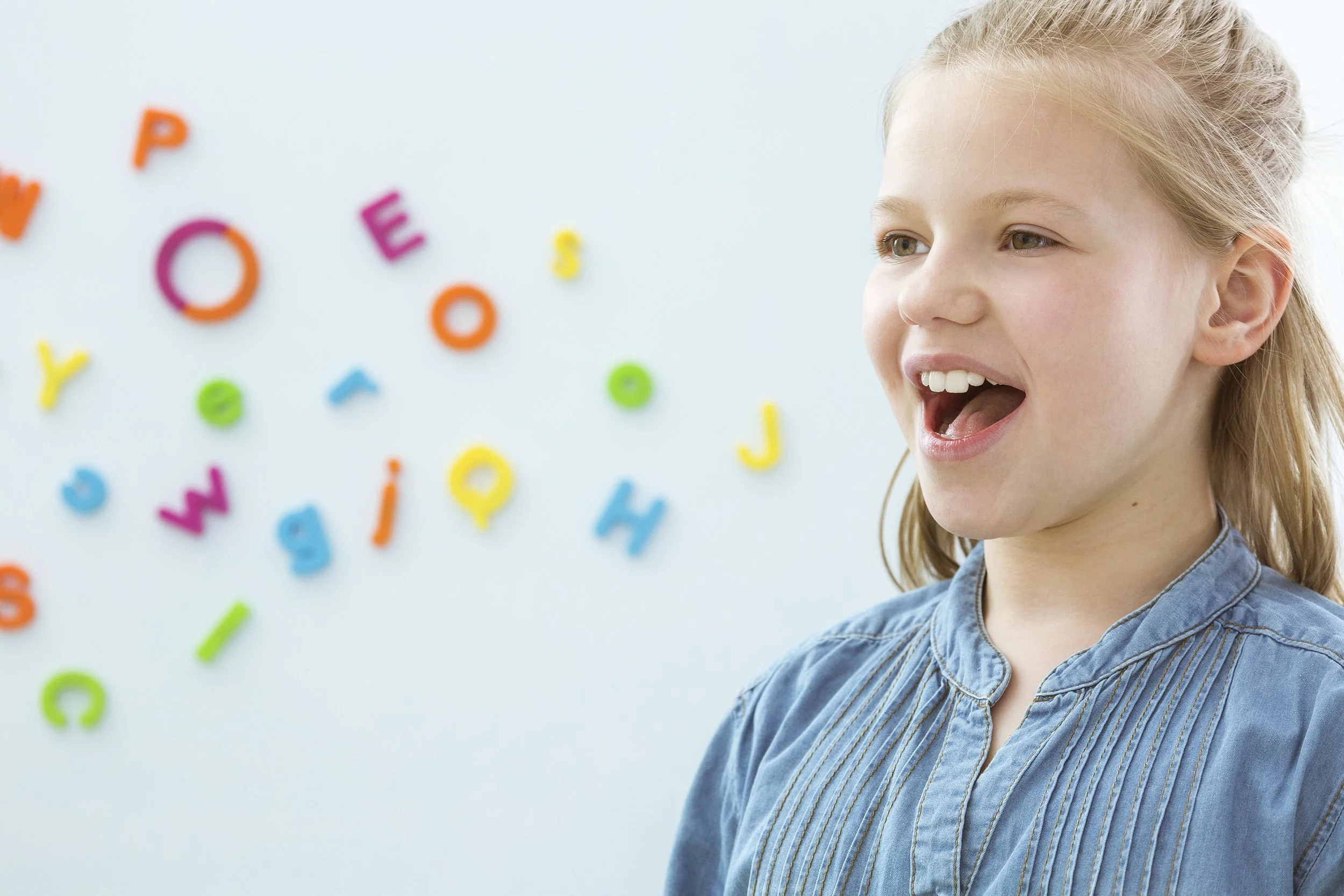 A young girl with blonde hair and a blue shirt smiling with her mouth open in front of a white wall with colorful magnetic alphabet letters.