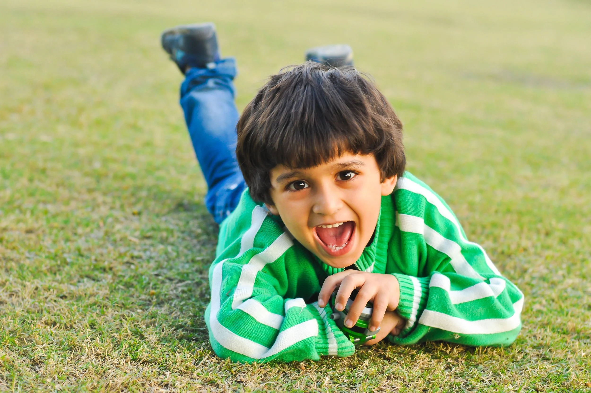 A young boy lying on the grass, smiling widely with his mouth open, wearing a green and white striped hoodie.