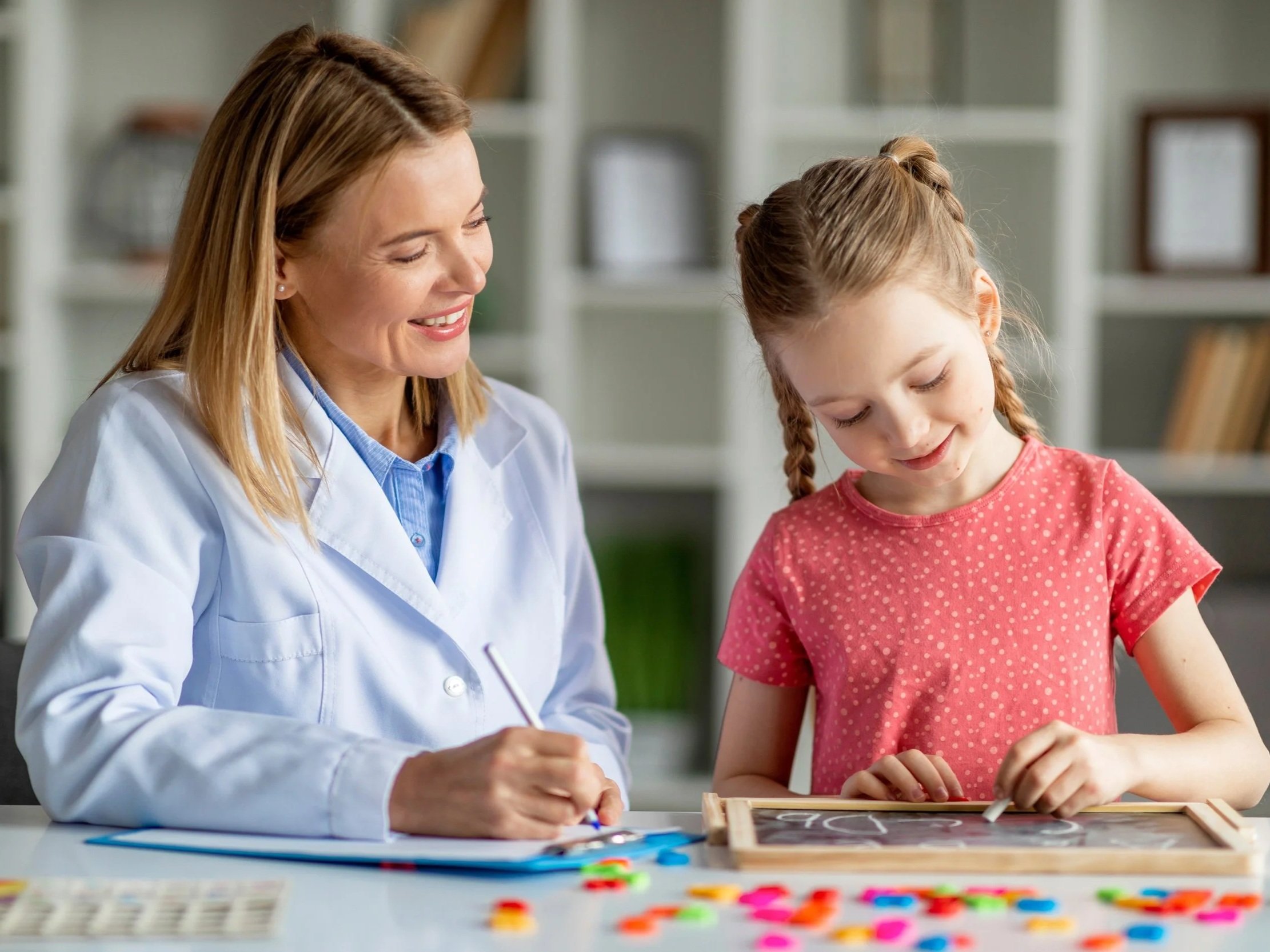 A woman and a young girl working together on a craft project at a table. The woman is smiling and the girl is focused on her task, with craft supplies such as paper and stickers on the table.