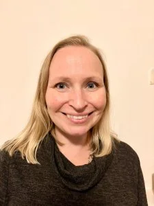 A woman with blonde hair smiling in front of a plain beige wall, wearing a dark top.