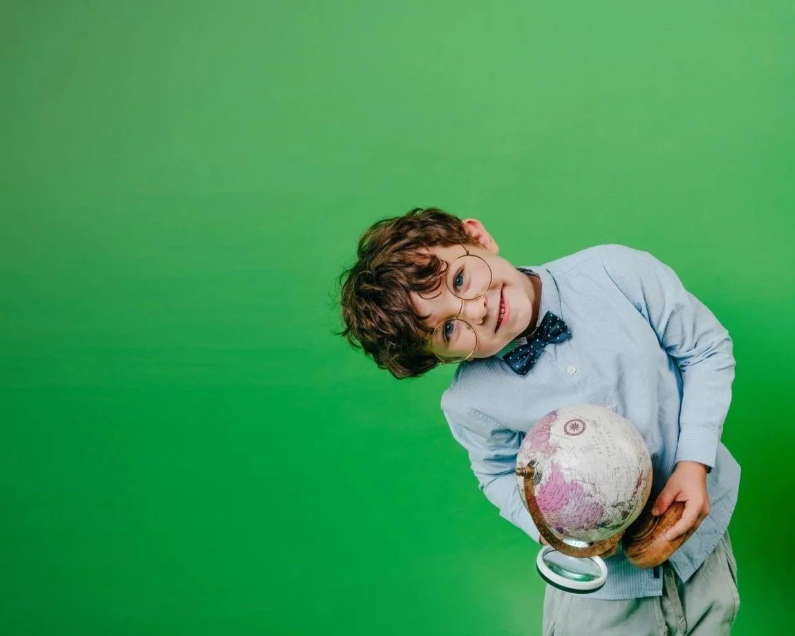 Young boy with curly hair and glasses, dressed in a light blue shirt and bow tie, holding a globe, standing against a green background