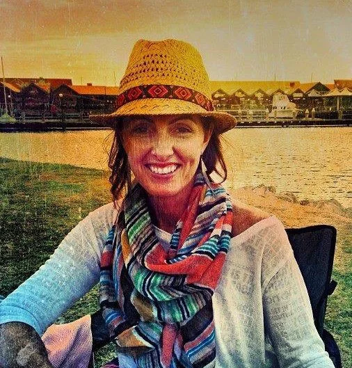 Woman smiling outdoors by a river at sunset, wearing a straw hat, colorful scarf, and light-colored shirt. Sherri Murphy