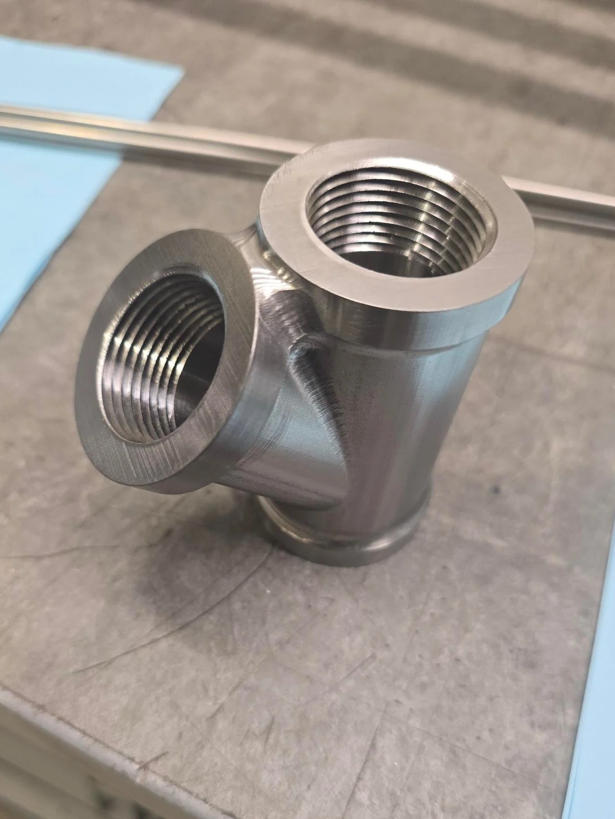 A metallic T-shaped pipe fitting with threaded openings on each end, placed on a grey surface.