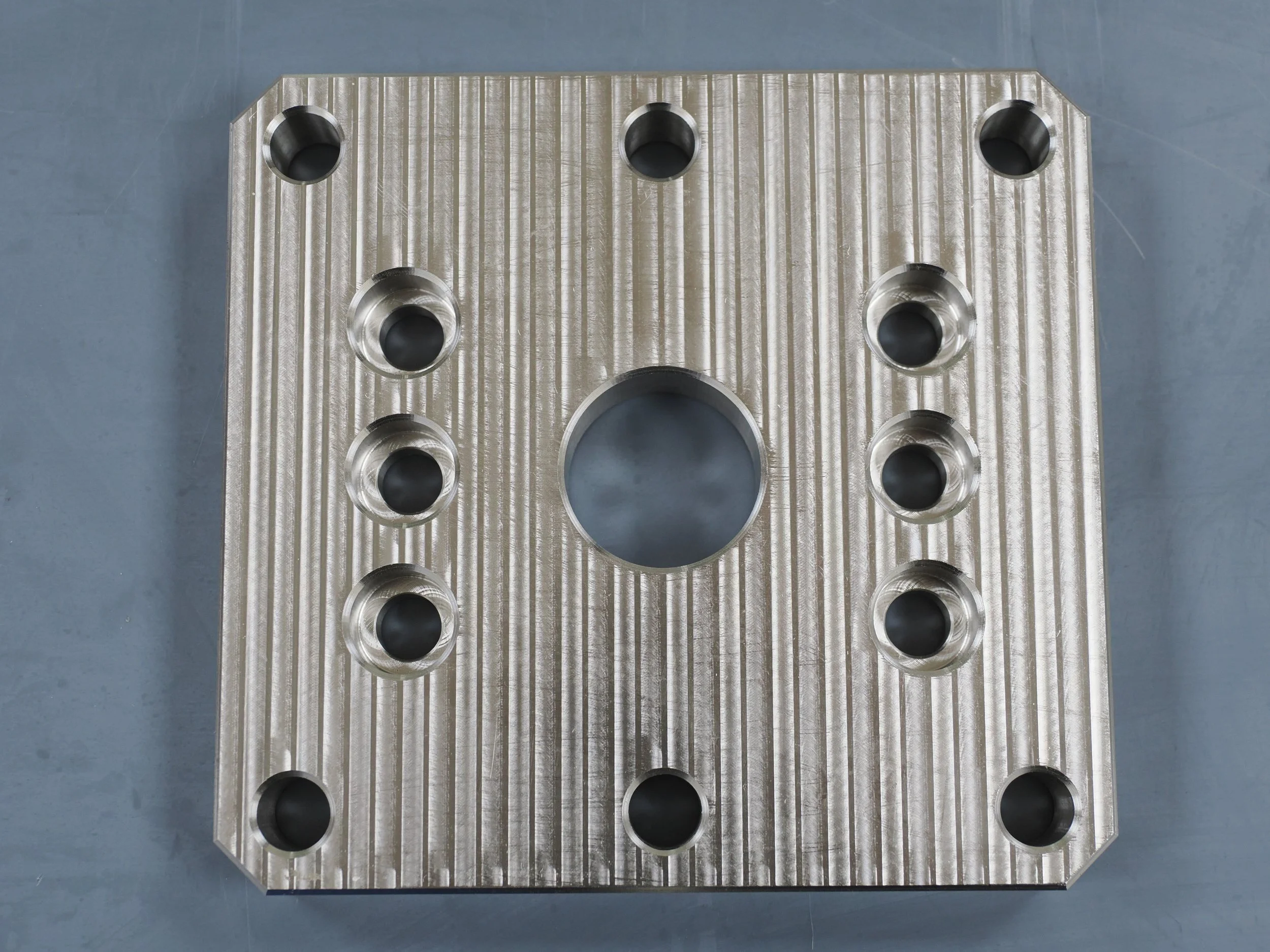 A metallic rectangular component with multiple circular holes evenly spaced on its surface, laid on a gray surface.