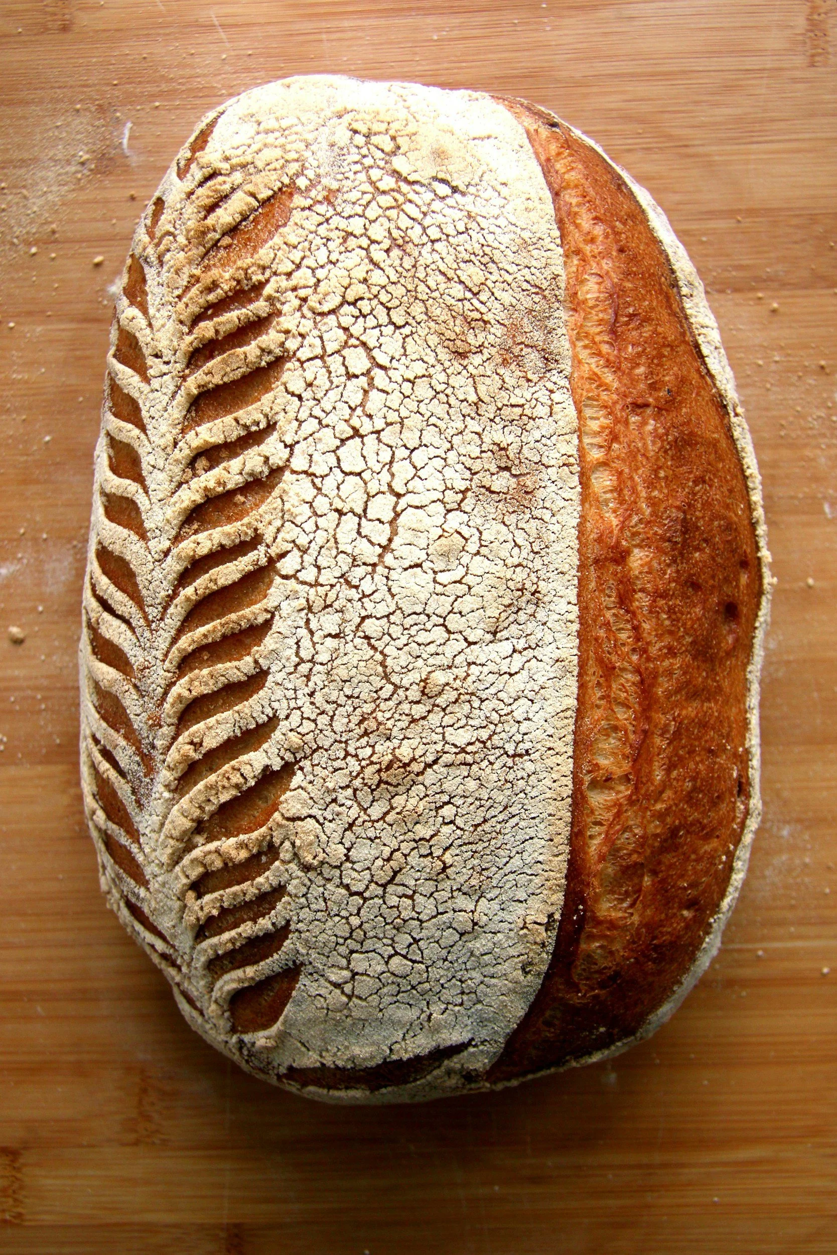 A loaf of bread with a decorative scoring pattern on top, dusted with flour, placed on a wooden surface.