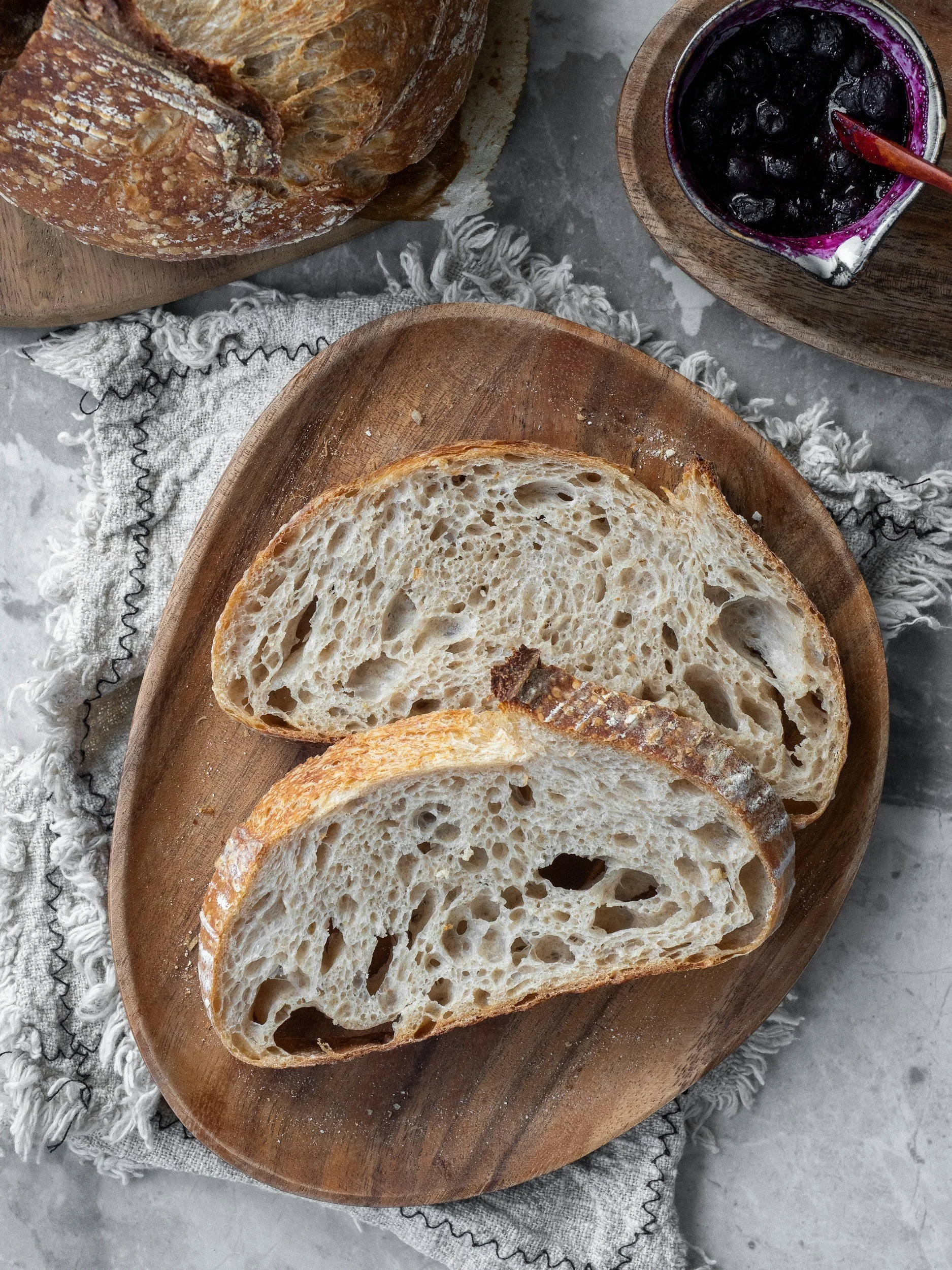 Sliced rustic bread on a wooden plate, with a loaf of bread and a jar of blueberry jam in the background on a gray marble surface.