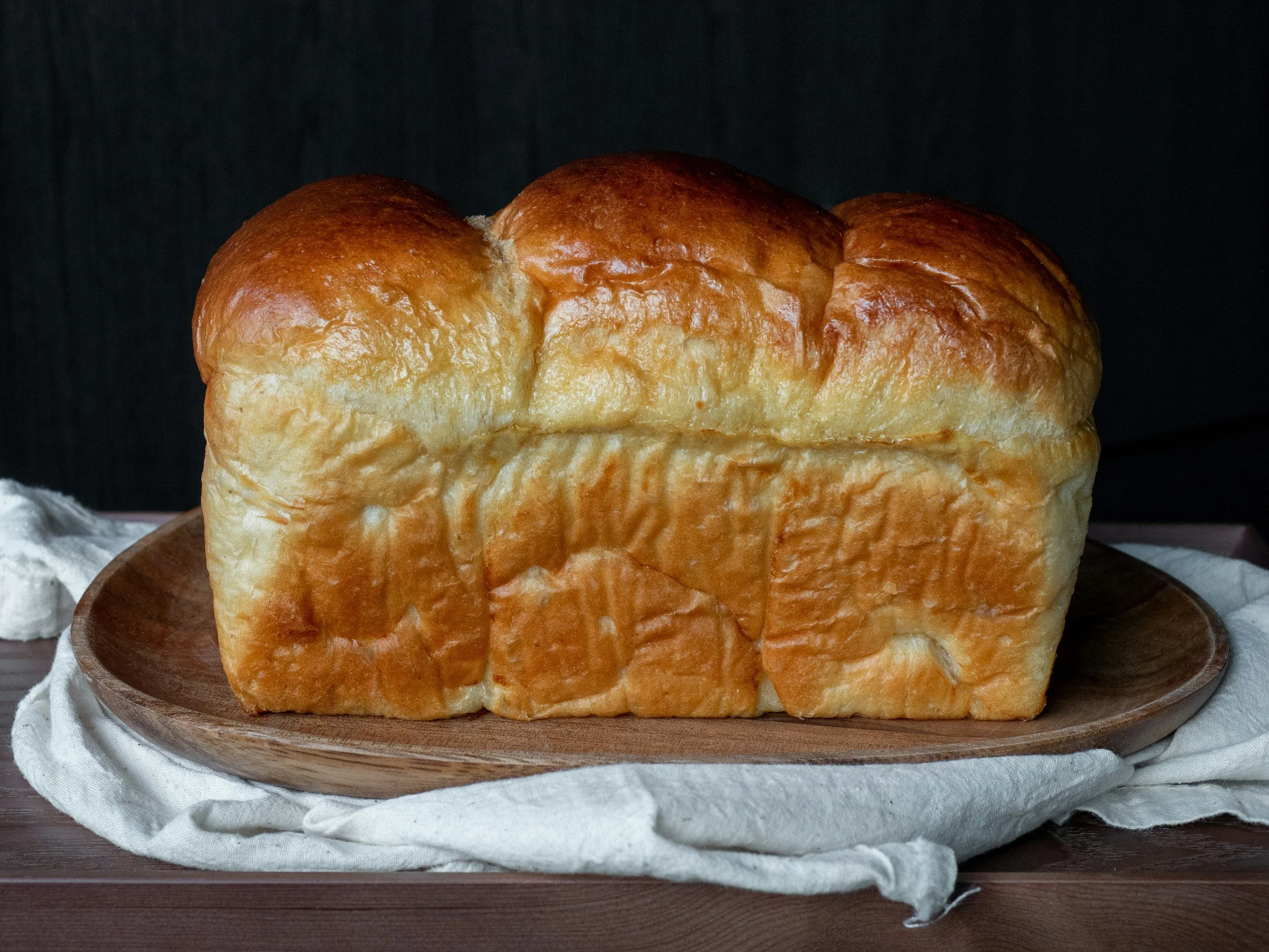 Loaf of freshly baked bread on a wooden plate with a white cloth underneath, dark background.