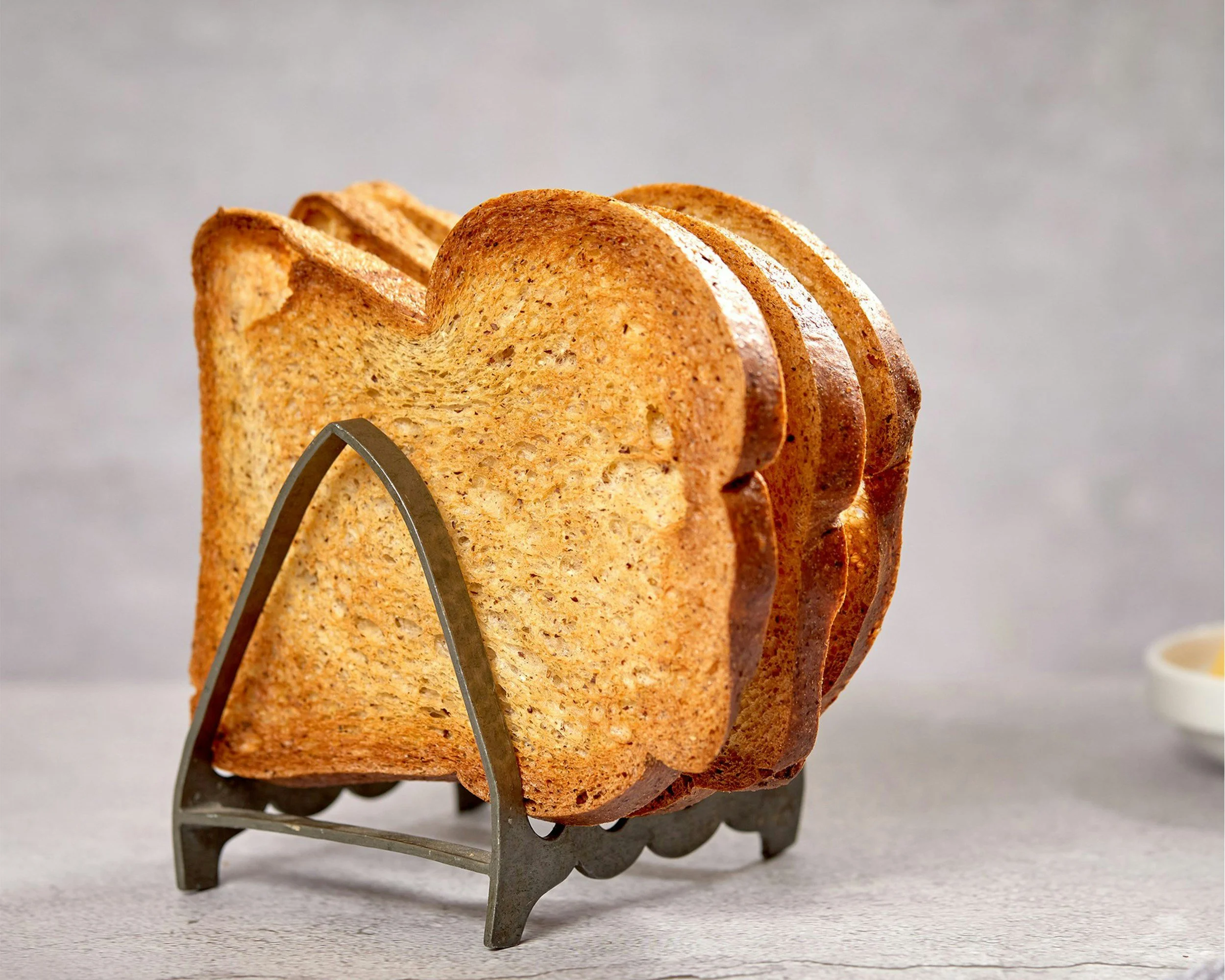 Several slices of toasted bread in a metal toast holder on a light surface with a small dish in the background.