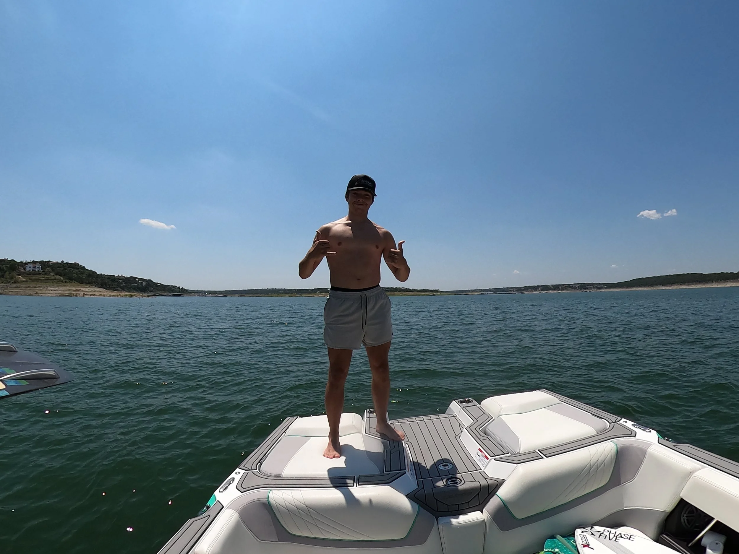 A young man standing shirtless on a boat, making a shaka sign with both hands, with water and distant shoreline in the background during a sunny day.