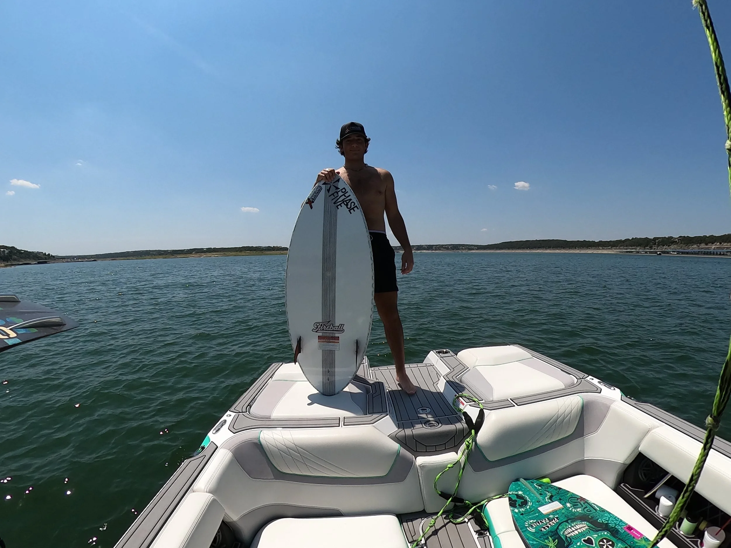 A young man in black shorts and a cap standing on a boat's deck holding a white surfboard with a gray stripe and red logo, on a sunny day with calm water and a distant shoreline.