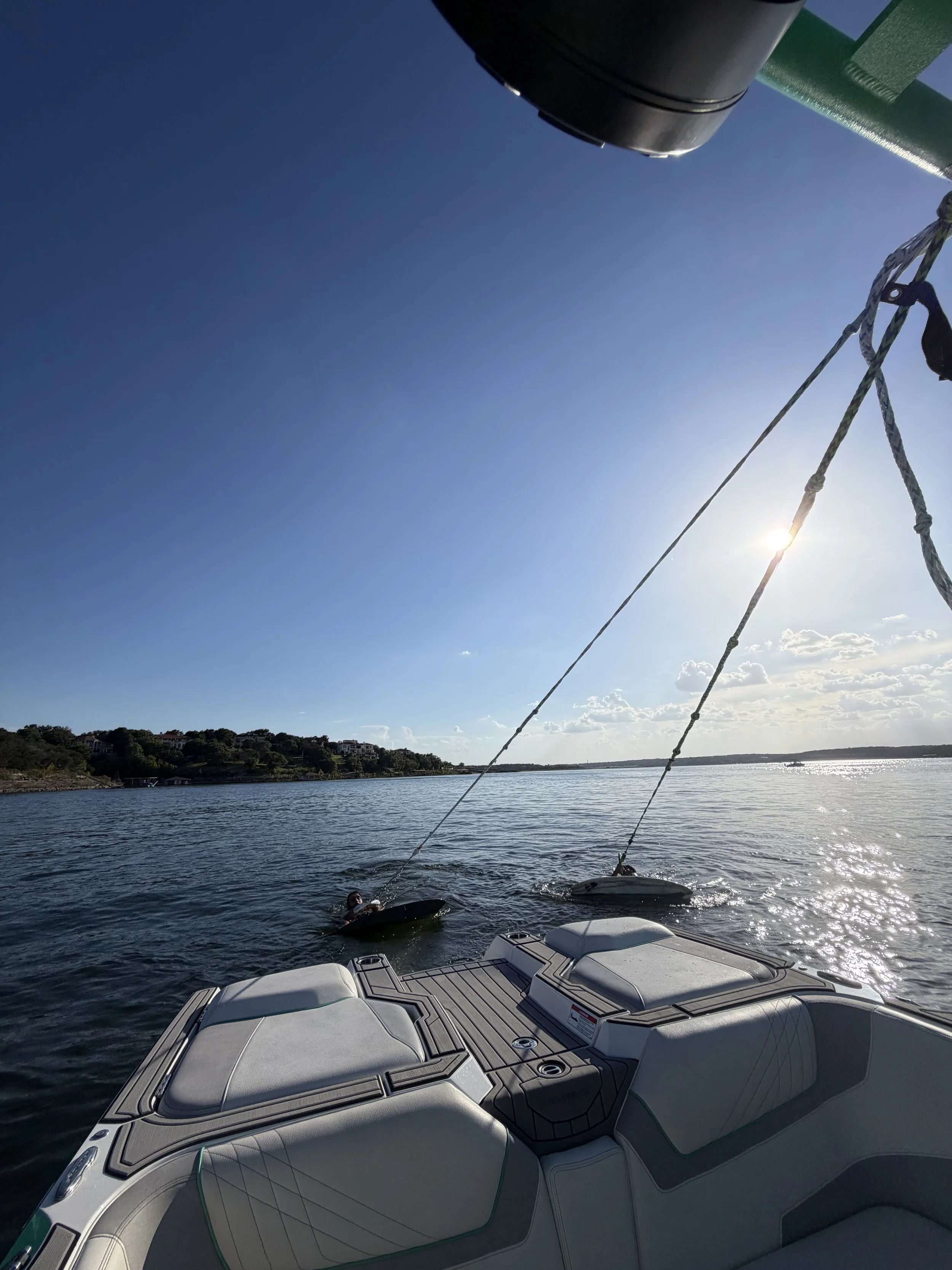 View from a boat showing a clear blue sky and a calm body of water with a shoreline in the distance and a boat tied to a mooring. Part of the boat's bow and deck with cushioned seating are visible in the foreground.