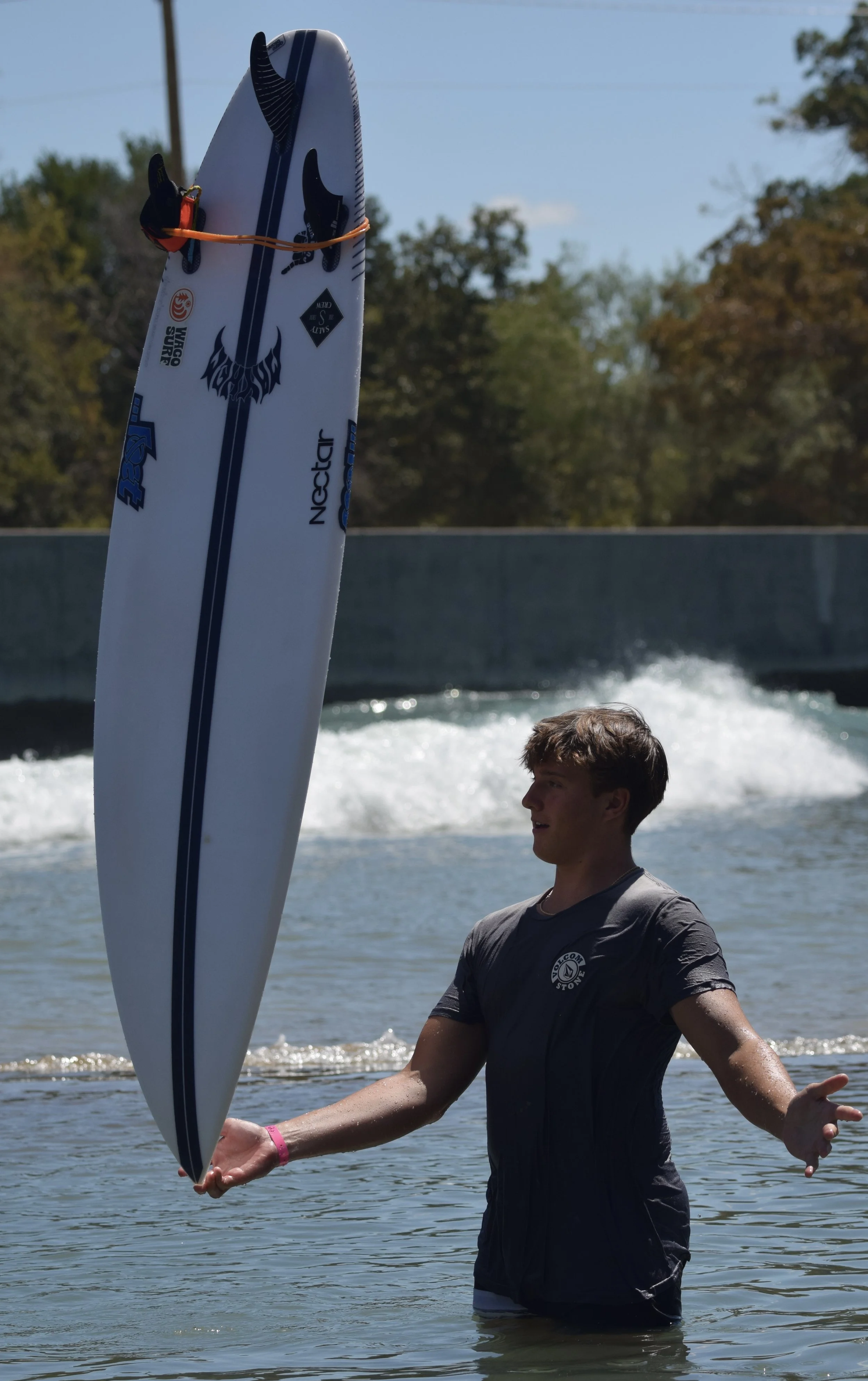 A young man in a wet t-shirt standing in shallow water, holding a white to blue paddleboard vertically in front of him. The sky is clear and there are trees in the background.