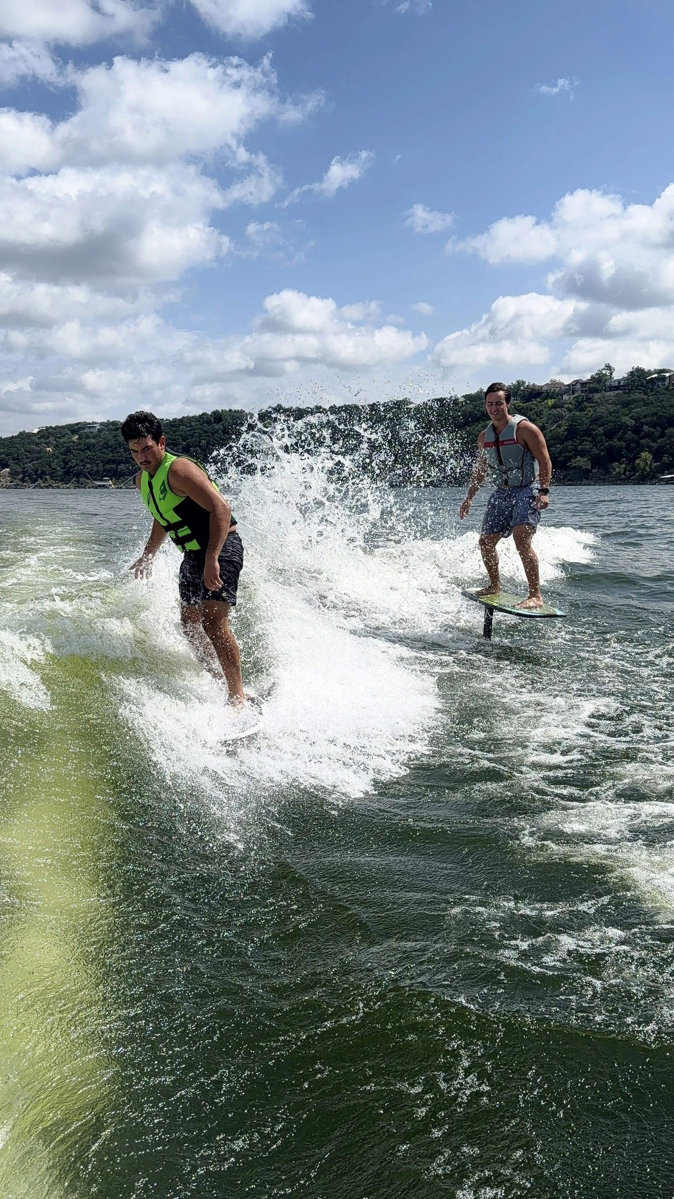 Two young men wakeboarding on a lake with a hill and houses in the background, under a partly cloudy sky.