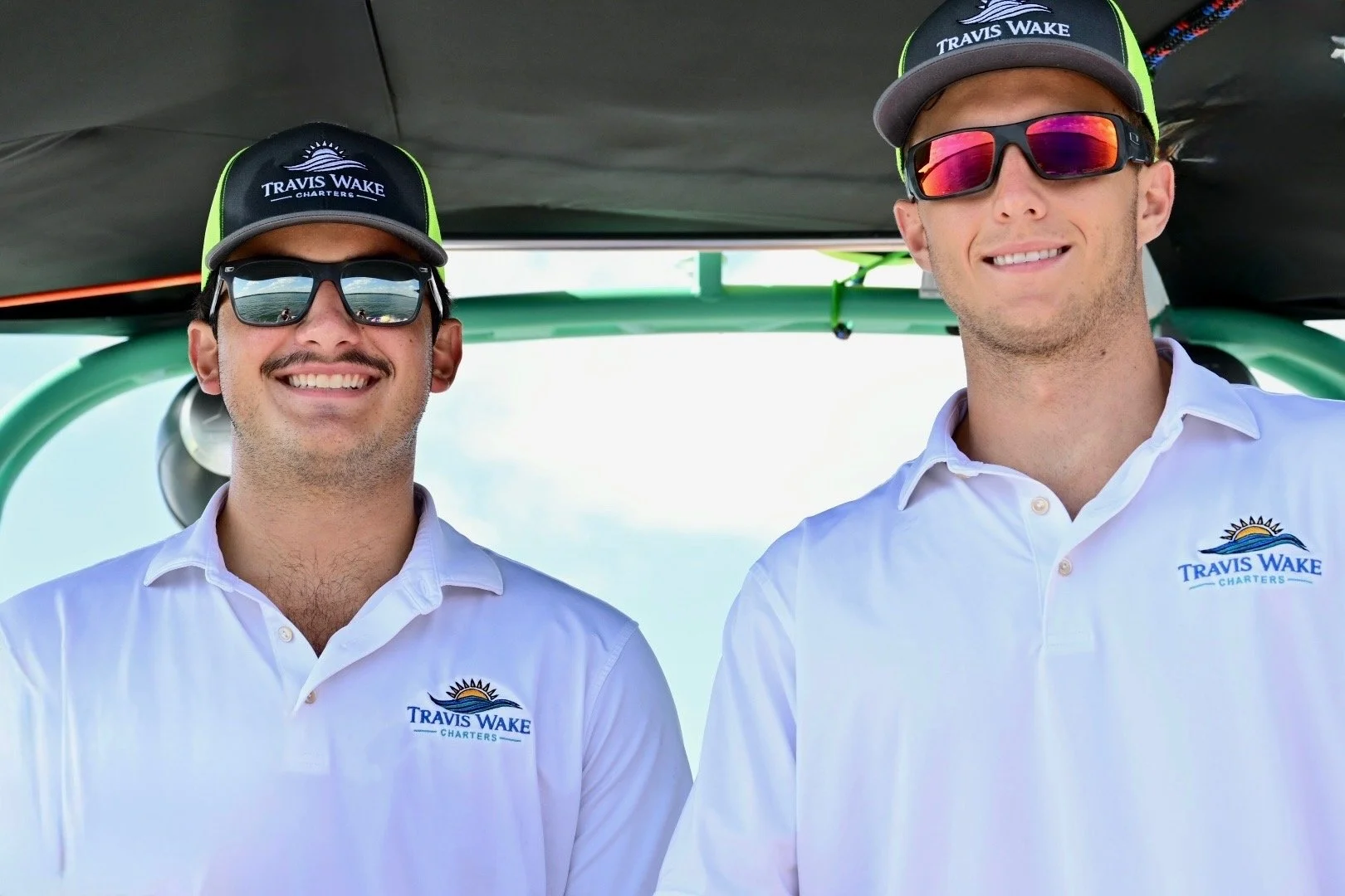 Two men wearing sunglasses and Travis Wake Charters hats, smiling and standing in front of a green and black boat, both in white polo shirts with the company's logo.