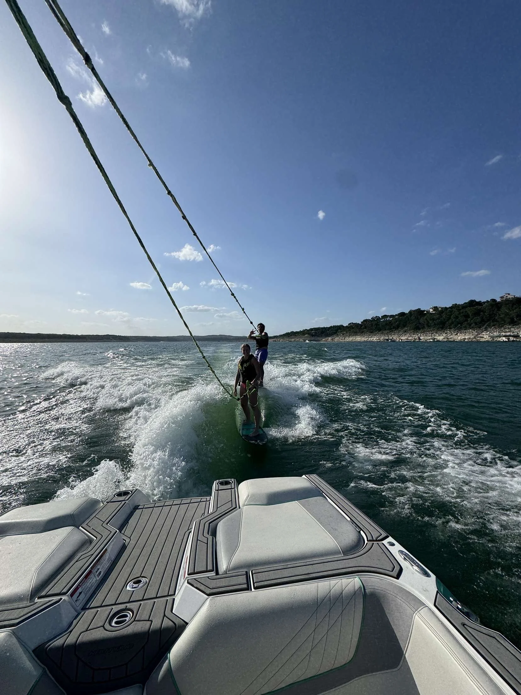 Two people wakeboarding behind a boat on a large body of water with a lake shoreline and trees in the distance, under a mostly clear blue sky.